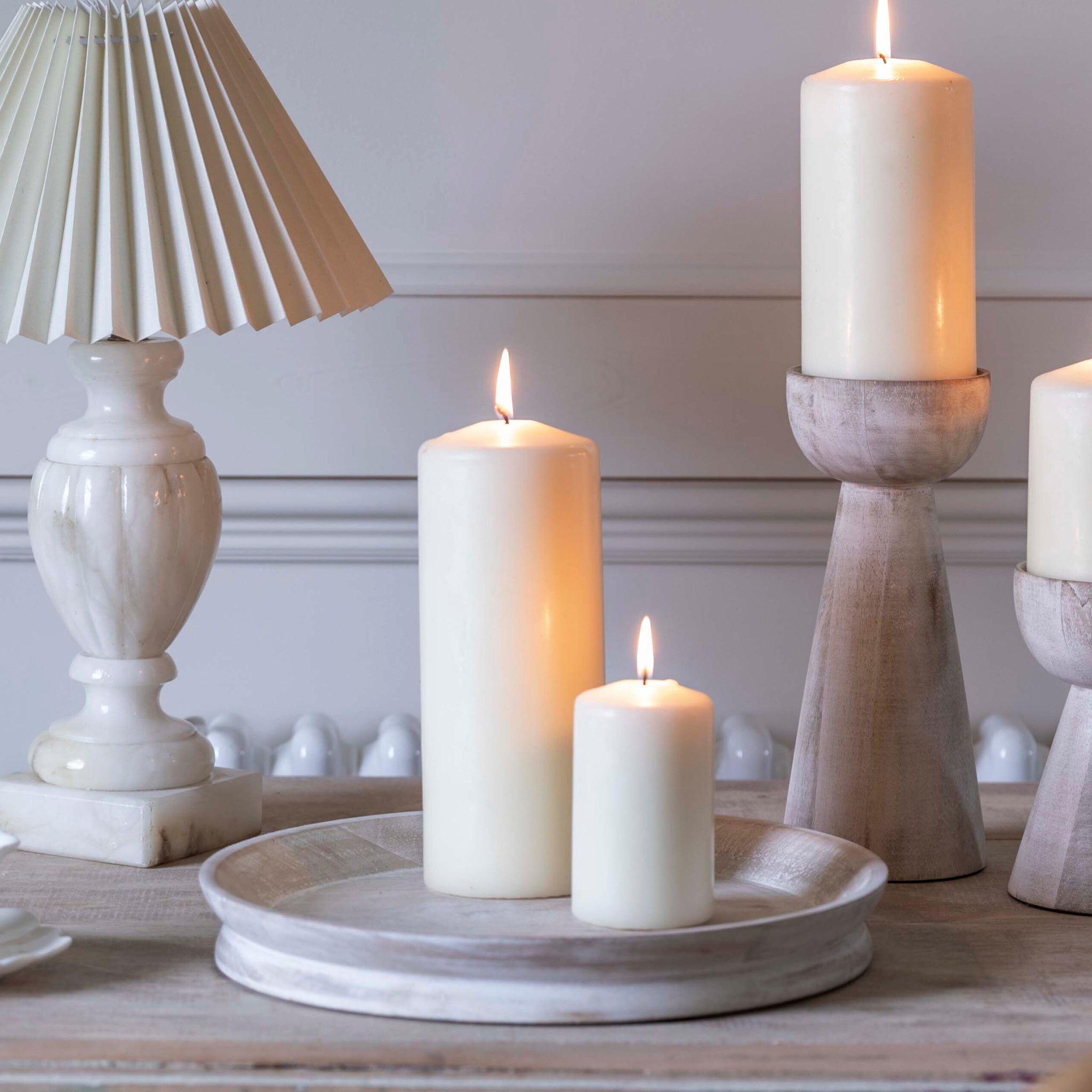 Lit white candles and a decorative lamp sit on an Ivyline Padstow Whitewash Wooden Candle Tray atop the wooden table in a softly lit room, blending seamlessly into the modern décor.