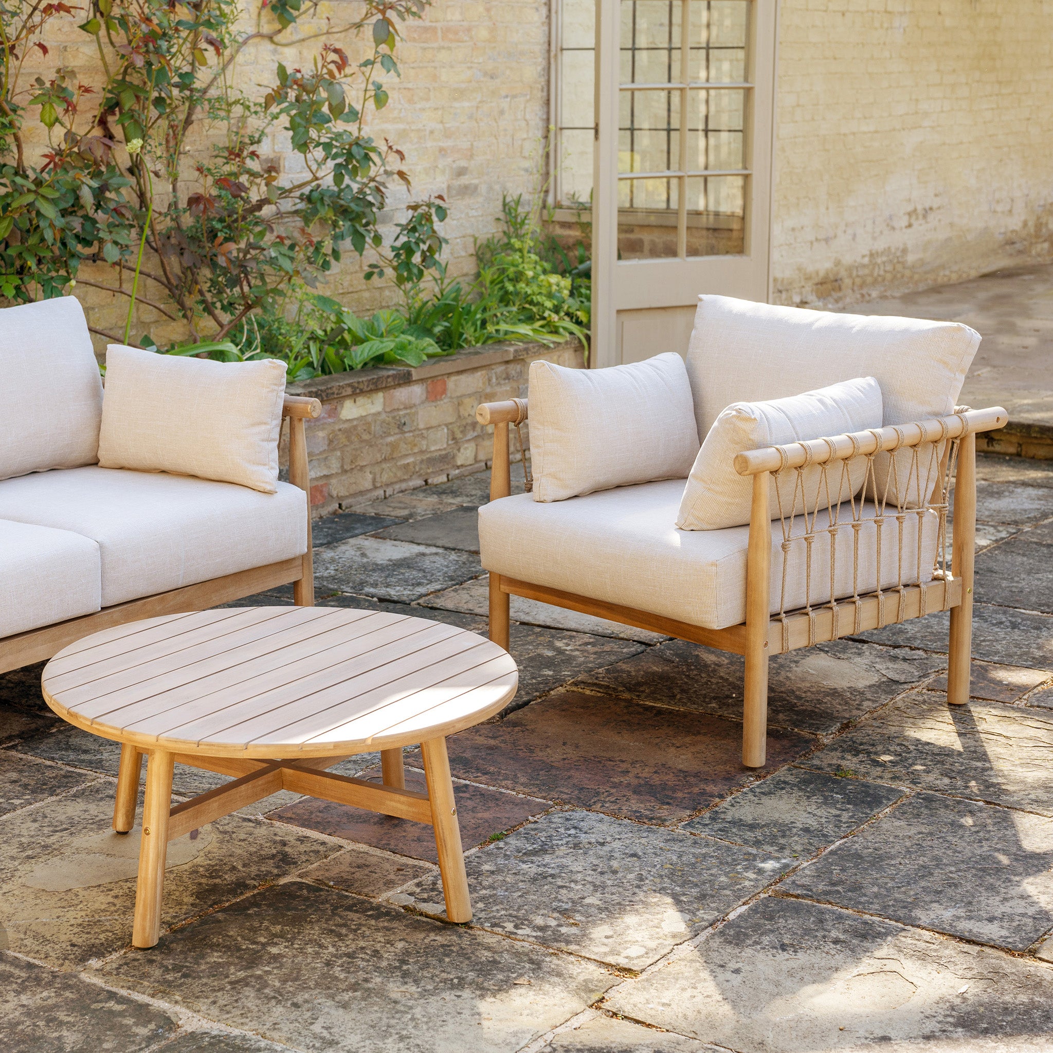 Paia 3 Seat Sofa Set in Linen with beige water-resistant polyester cushions, wooden table, and loveseat on a stone-tiled patio near a brick wall offers elegance and lasting durability for your garden.