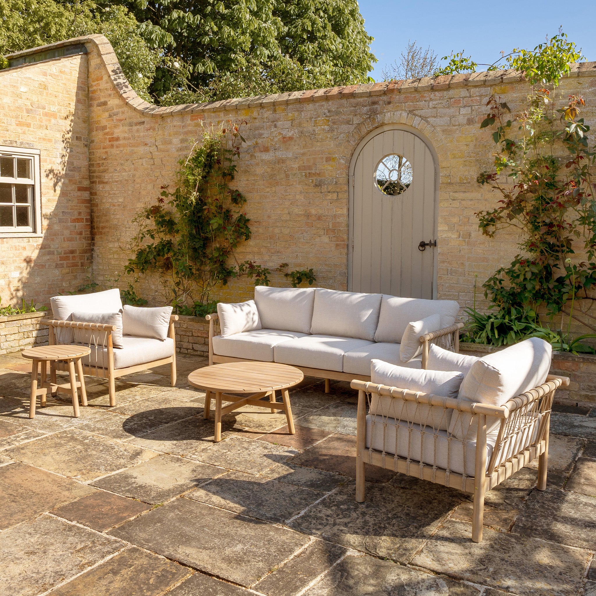 A patio with light wooden garden furniture from the Paia 3 Seat Sofa Set in Linen, including a sofa, two chairs, and two round tables made from water-resistant polyester, set against a brick wall and greenery backdrop.