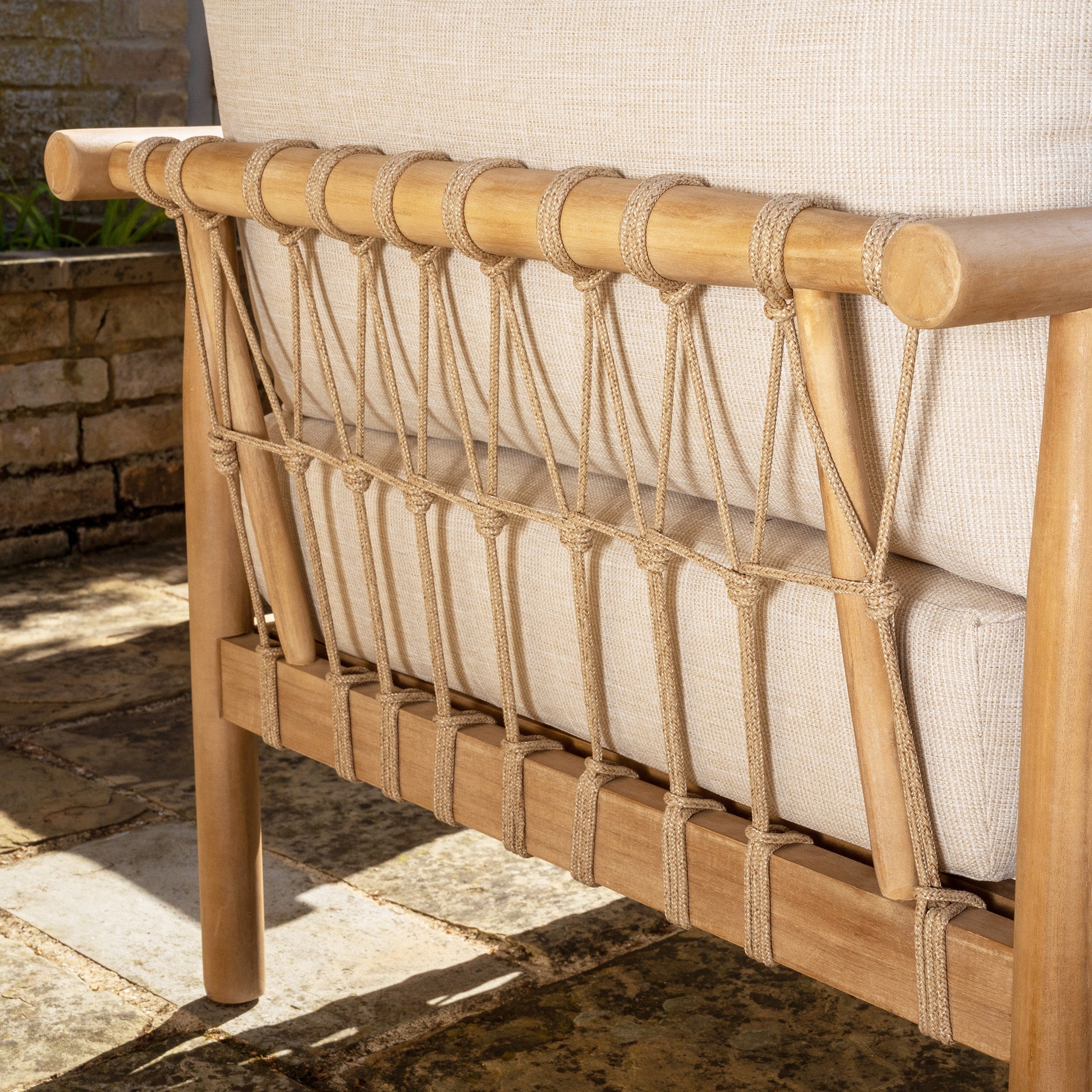 Close-up of the Paia 3 Seat Sofa Set in Linen, featuring a beige outdoor chair with a woven backrest and wooden frame on a stone patio, showcasing its water-resistant polyester fabric ideal for garden furniture.