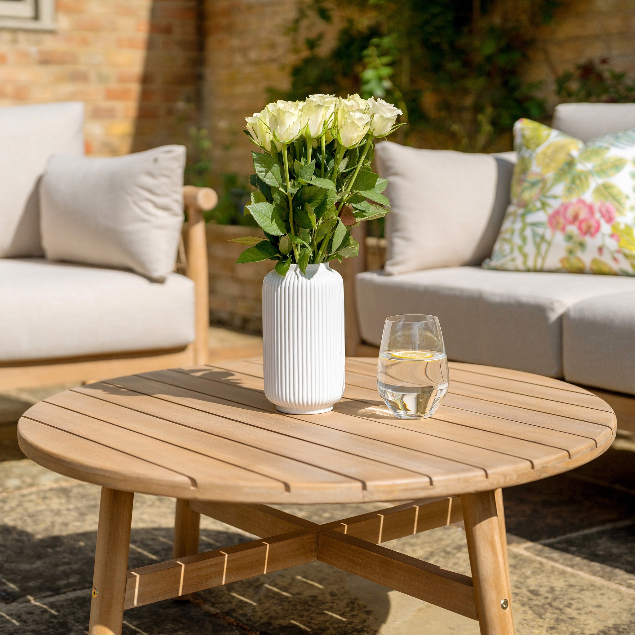 A Paia 3 Seat Sofa Set in Linen, featuring water-resistant polyester cushions, arranged in an outdoor seating area with a wooden table adorned with a white vase of flowers and a glass of water.