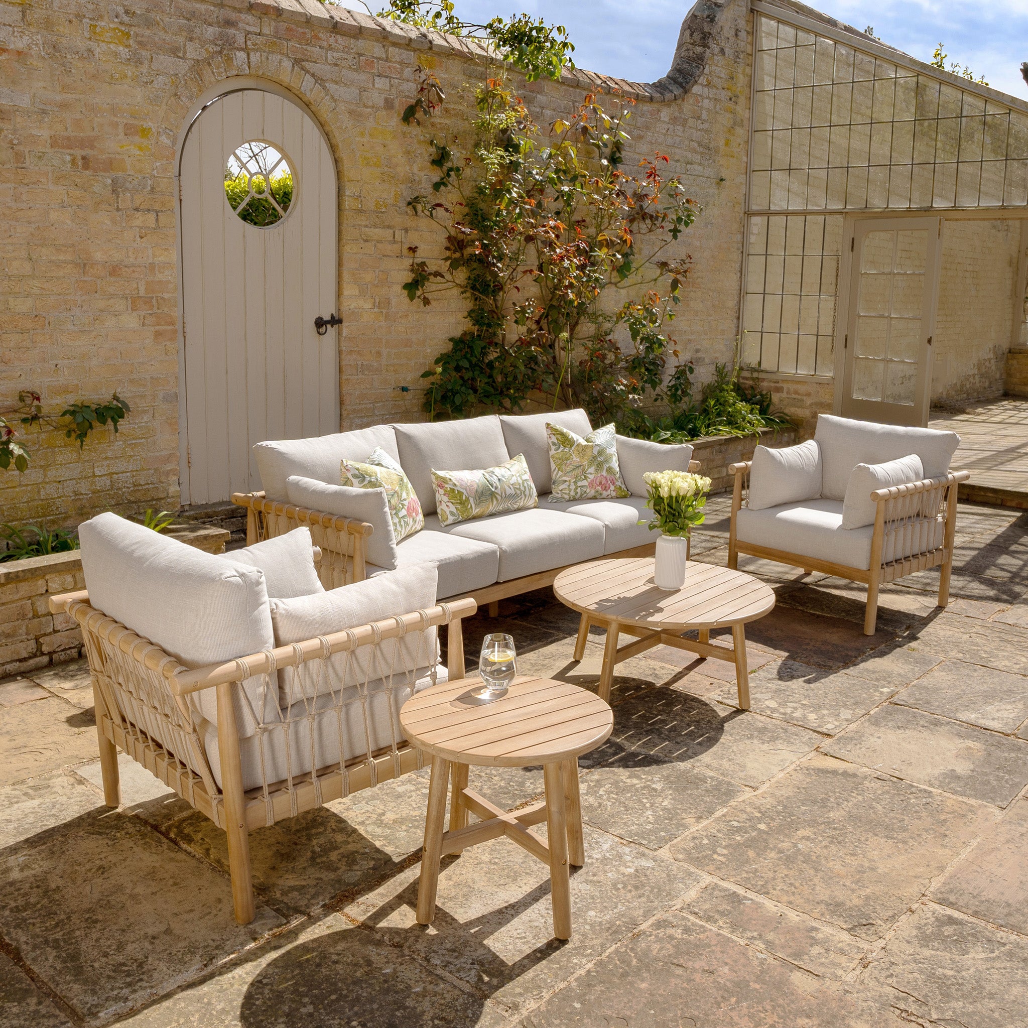 A cozy outdoor seating setup featuring the Paia 3 Seat Sofa Set in Linen, complemented by beige woven garden furniture, two round wooden tables, and potted plants against a brick wall with a door.