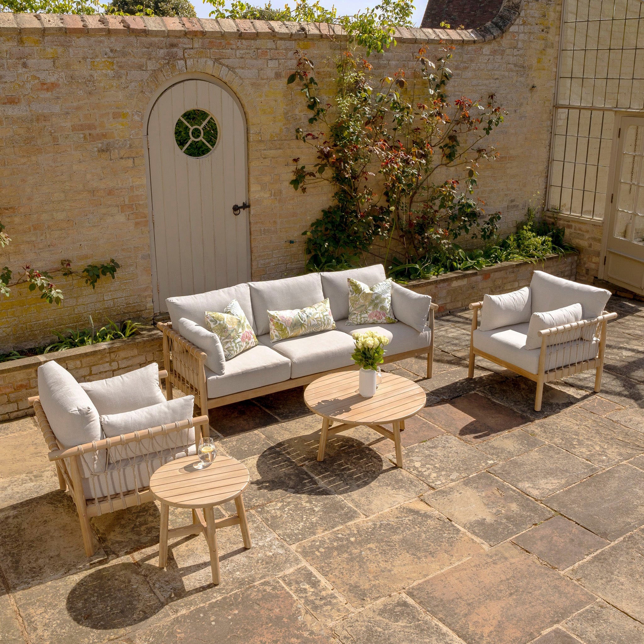 Enjoy a cozy outdoor patio with the Paia 3 Seat Sofa Set in Linen, complemented by cushioned wooden furniture, round tables, and floral decor. The inviting space is set against a brick wall with a door. The garden furniture features water-resistant polyester cushions, ensuring lasting comfort for your outdoor seating experience.