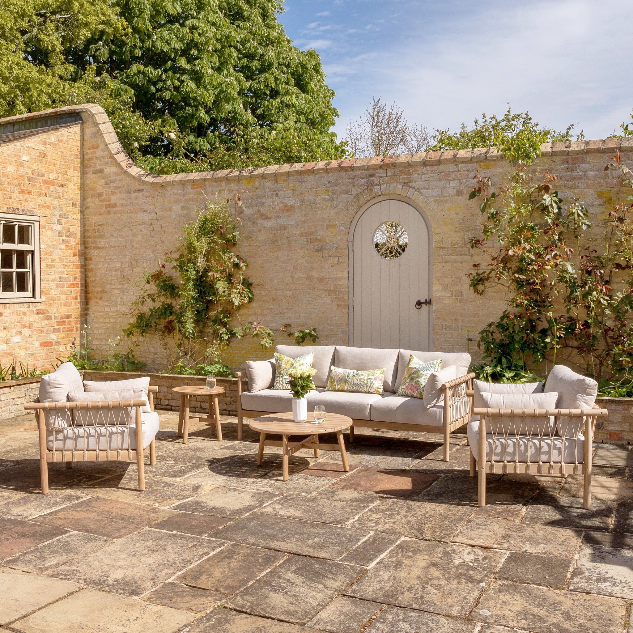 Outdoor patio featuring the Paia 3 Seat Sofa Set in Linen, along with matching armchairs, coffee tables, and water-resistant polyester cushions, all set against a brick wall with potted plants enhancing the ambiance.