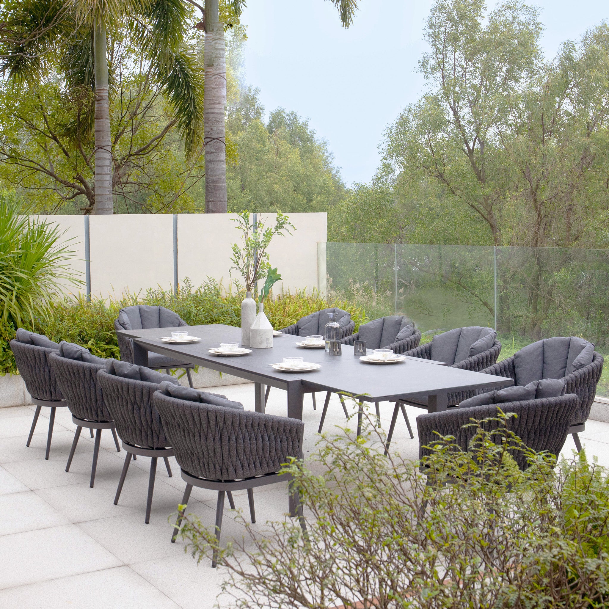 Outdoor dining area featuring the Palma 10 Seat Rope Extending Dining Set in Grey, complete with aluminium frames, a long table, ten chairs, place settings, and decorative items, all surrounded by greenery and a glass fence.