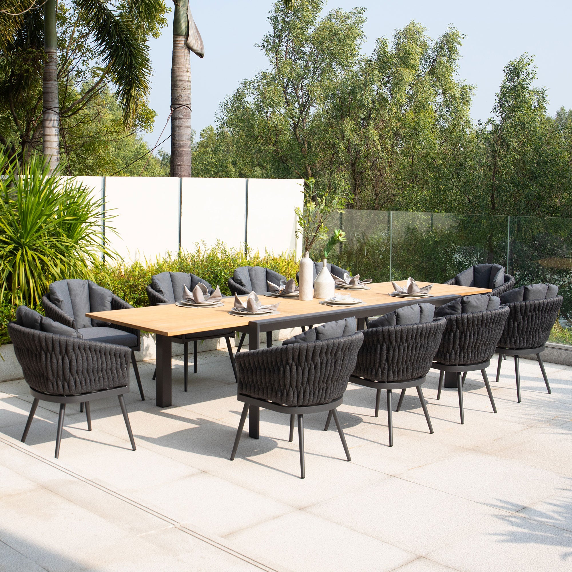 The Harbour Lifestyle Palma 10 Seat Rope Extending Teak Dining Set in Grey features a long teak table, ten black cushioned chairs, and vibrant greenery in the background.