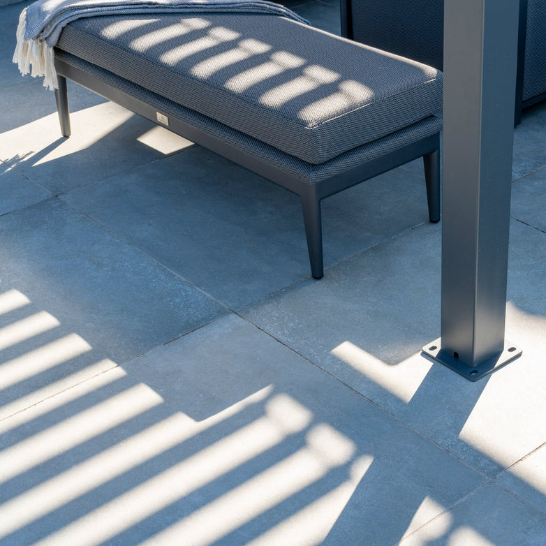 An outdoor bench on a patio, covered with a blanket. Shadows of the PergoSTET 3m x 3m Metal Square Pergola in Grey's tilting louvres cast patterns on the ground.