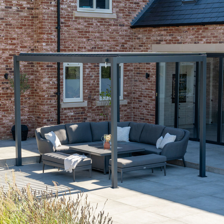 Modern outdoor patio featuring a gray sectional sofa and the sleek PergoSTET 3m x 4m Metal Rectangular Pergola in Grey with tilting louvres on the tiled ground, attached to a brick house with large windows. The PergoSTET pergola comes with a waterproof roof for all-weather enjoyment.