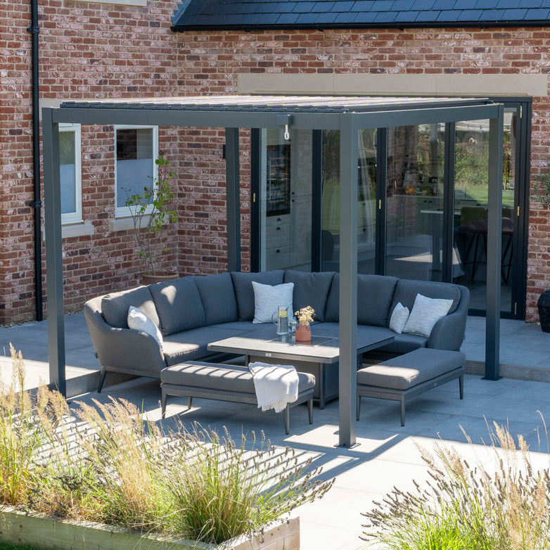 Outdoor patio with grey sectional sofas under a PergoSTET 3m x 3m Metal Square Pergola in Grey featuring tilting louvres, surrounded by potted plants in a garden setting.