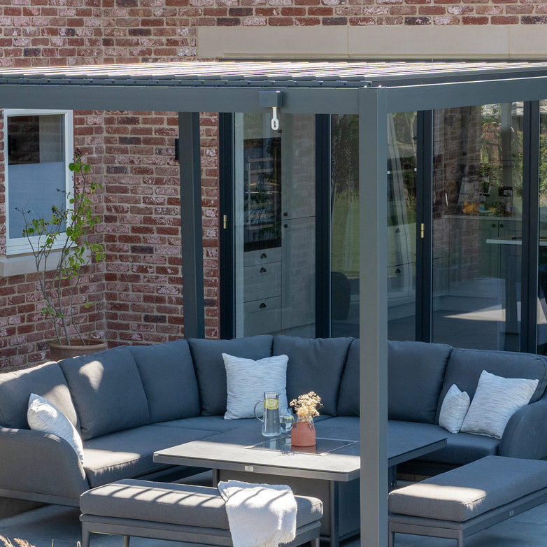 Outdoor patio with a gray sectional sofa, coffee table, and a PergoSTET 3m x 4m Metal Rectangular Pergola in Grey featuring tilting louvres and a waterproof roof against a brick house with large glass doors.