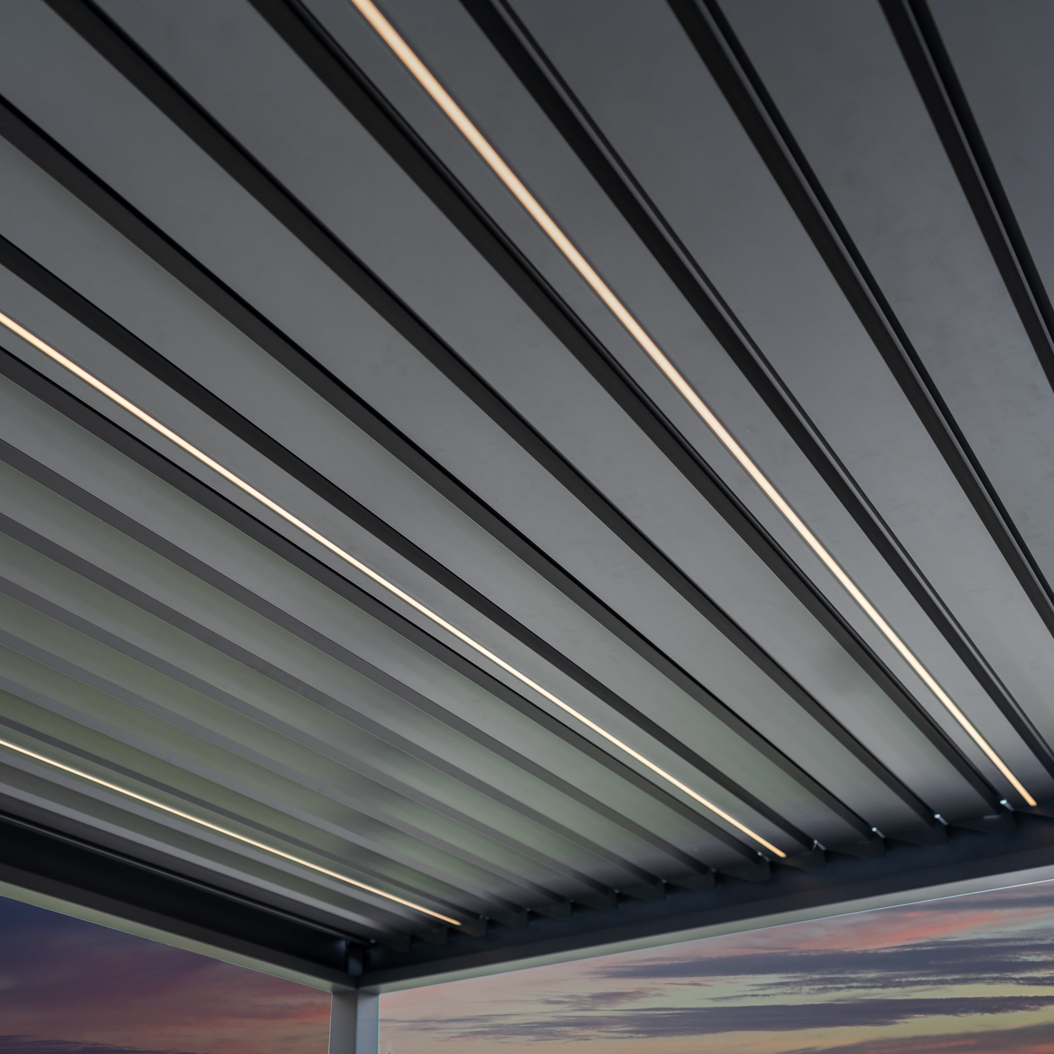 The PergoSTET Deluxe 3m x 9m rectangular pergola in grey features motorized LED louvres and a modern metal slatted design, perfectly complementing a colorful sunset sky.