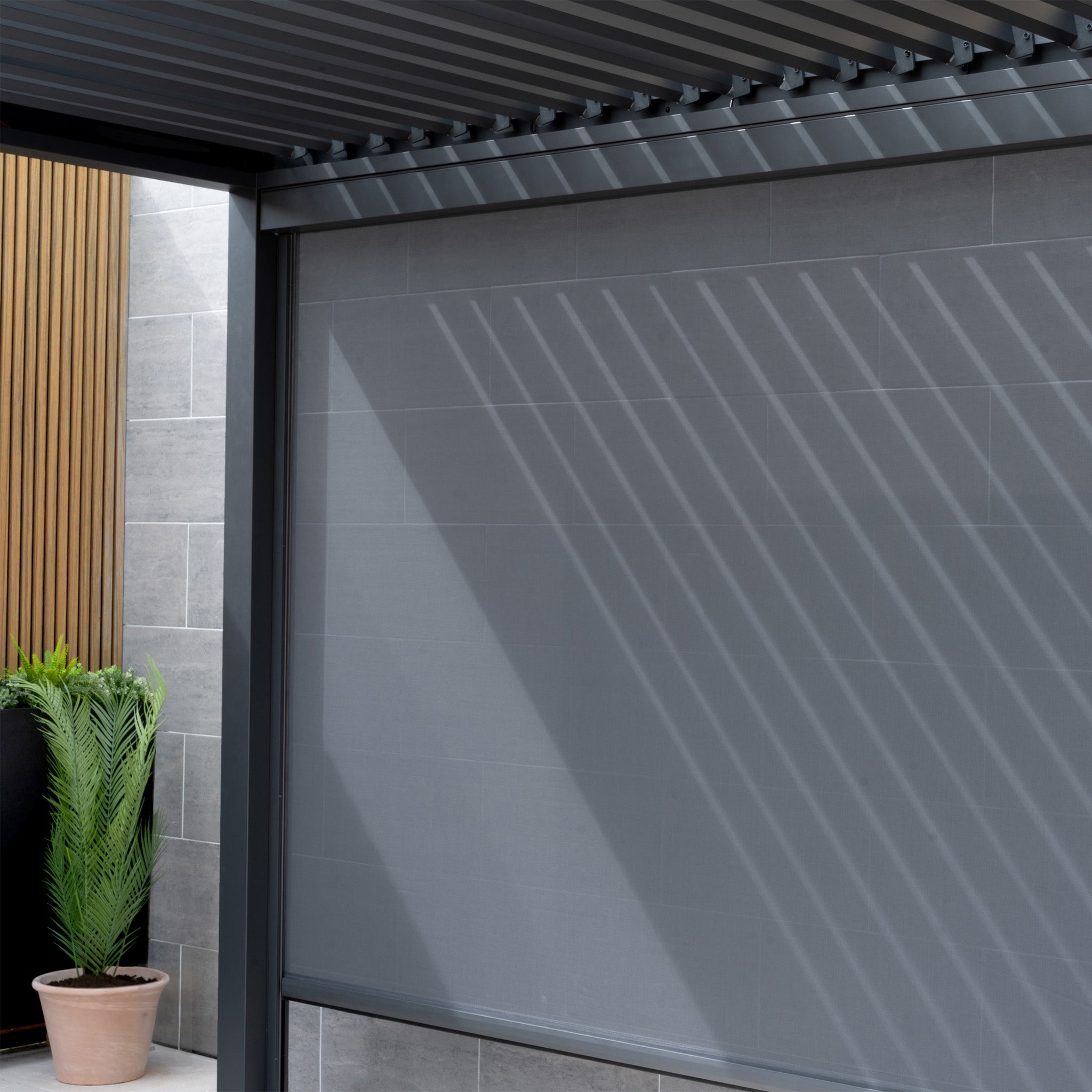 A modern patio with a PergoSTET Deluxe Manual Side Blind 3m in Grey creates striped shadows on a gray wall, complemented by potted plants.