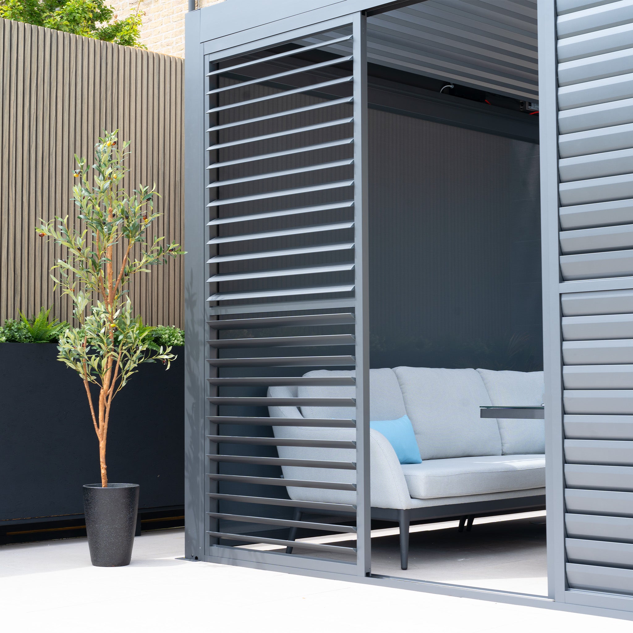 A modern outdoor setup features the PergoSTET Deluxe Louvred Sliding Doors in grey, paired with a white sofa, blue pillow, and a potted tree on a sunny patio.