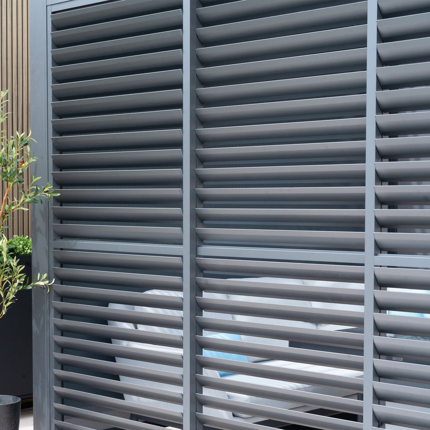 The PergoSTET Deluxe Louvred Side Panel for 3m Side in Grey features horizontal slats, partially shading an outdoor space. A plant peeks through the elegant slatted aluminum structure.