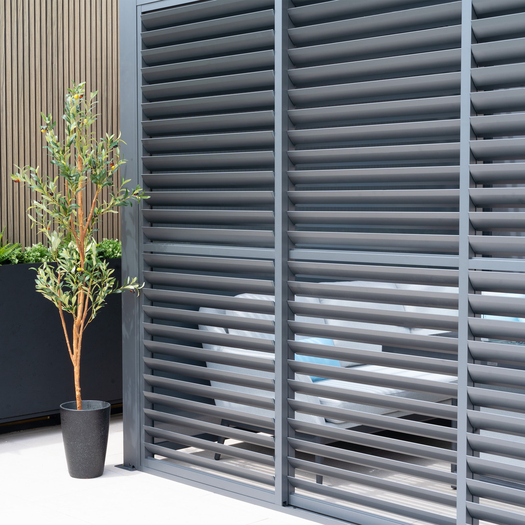 Potted plant beside the PergoSTET Deluxe Louvred Sliding Doors 3m Side in Grey, revealing glimpses of outdoor seating.