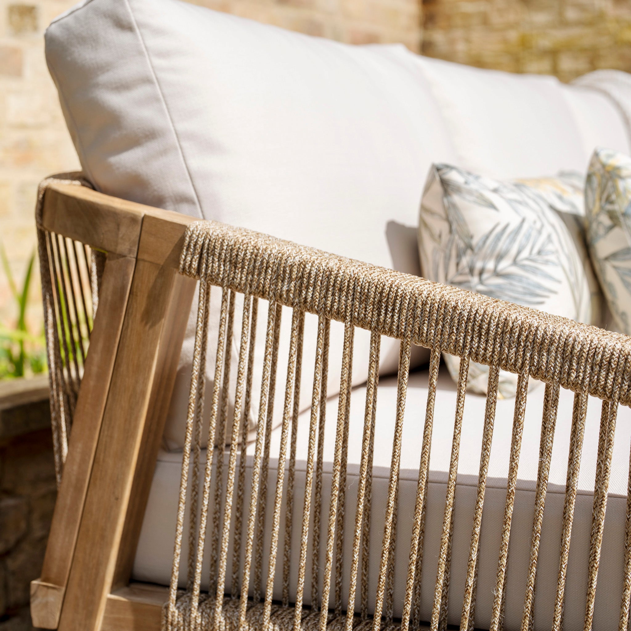 Close-up of a Quay Corner Dining Set with Armchair in Linen, showcasing a wicker outdoor arrangement with white cushions and floral pillows, set against a stone wall in a sunlit garden. Crafted from sustainably sourced acacia wood.