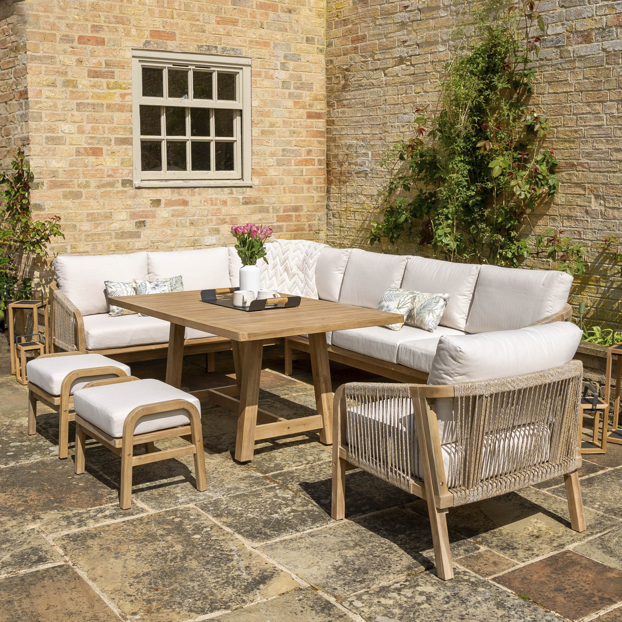 Outdoor patio with a Quay Corner Dining Set with Armchair in Linen made from sustainably sourced acacia wood, featuring white cushions, a table, two ottomans, and potted flowers, set against a brick wall.