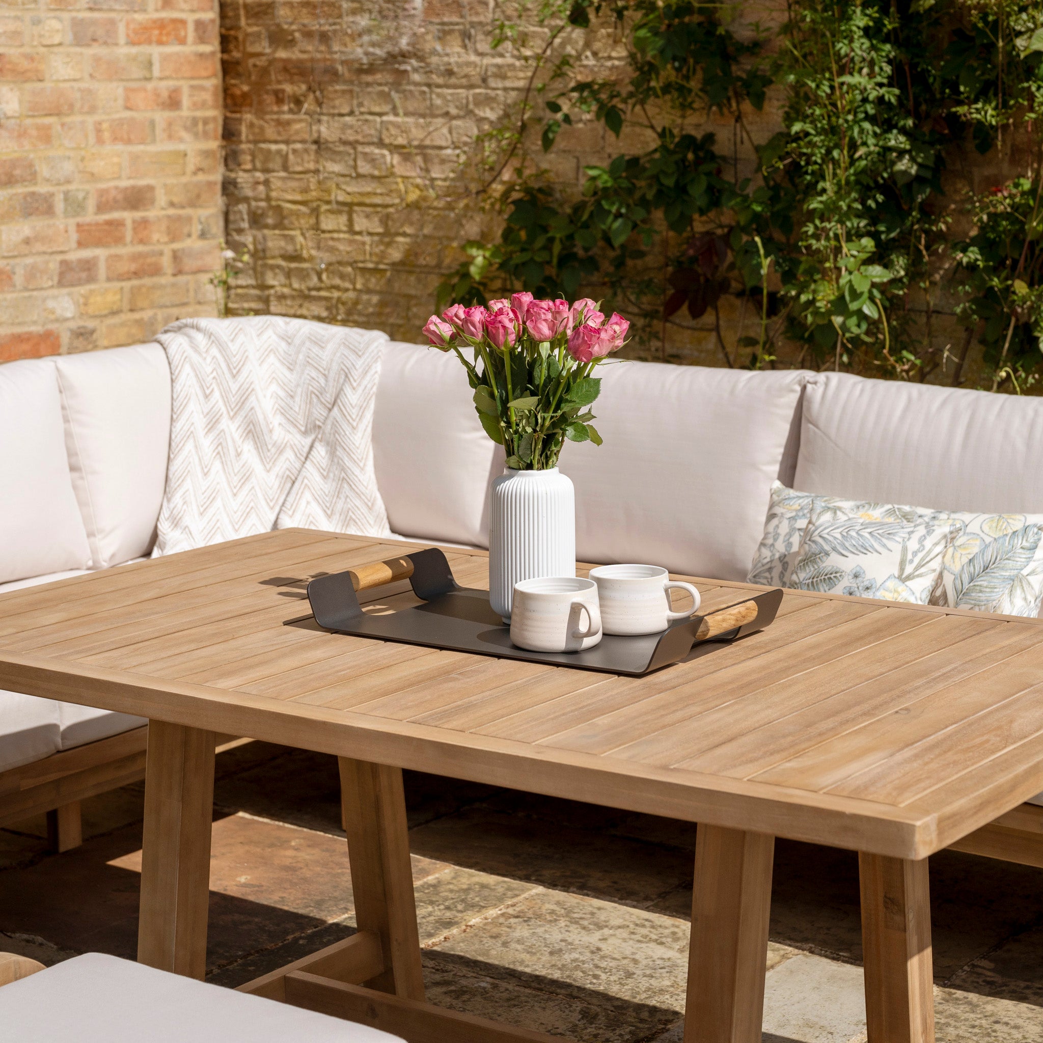 A wooden outdoor table crafted from sustainably sourced acacia wood, adorned with a vase of pink flowers, a tray with a mug and teapot, and paired with a white cushioned bench. Perfect for enhancing your outdoor furniture setup or complementing the Quay Corner Dining Set with Armchair in Linen.