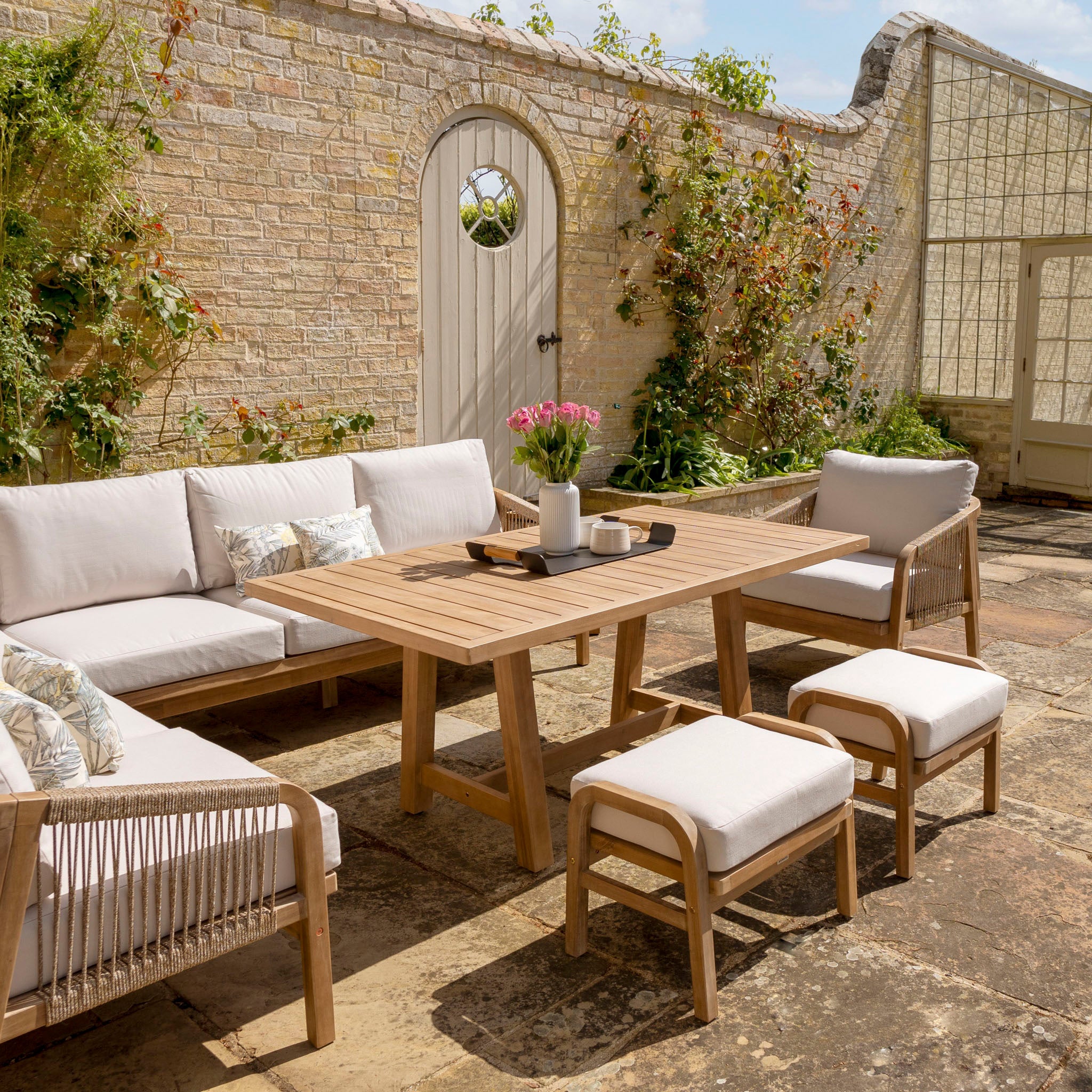 A cozy patio featuring the Quay Corner Dining Set with Armchair in Linen, crafted from sustainably sourced acacia wood, with white cushions and blooming flowers, set against a stone wall with an arched doorway.