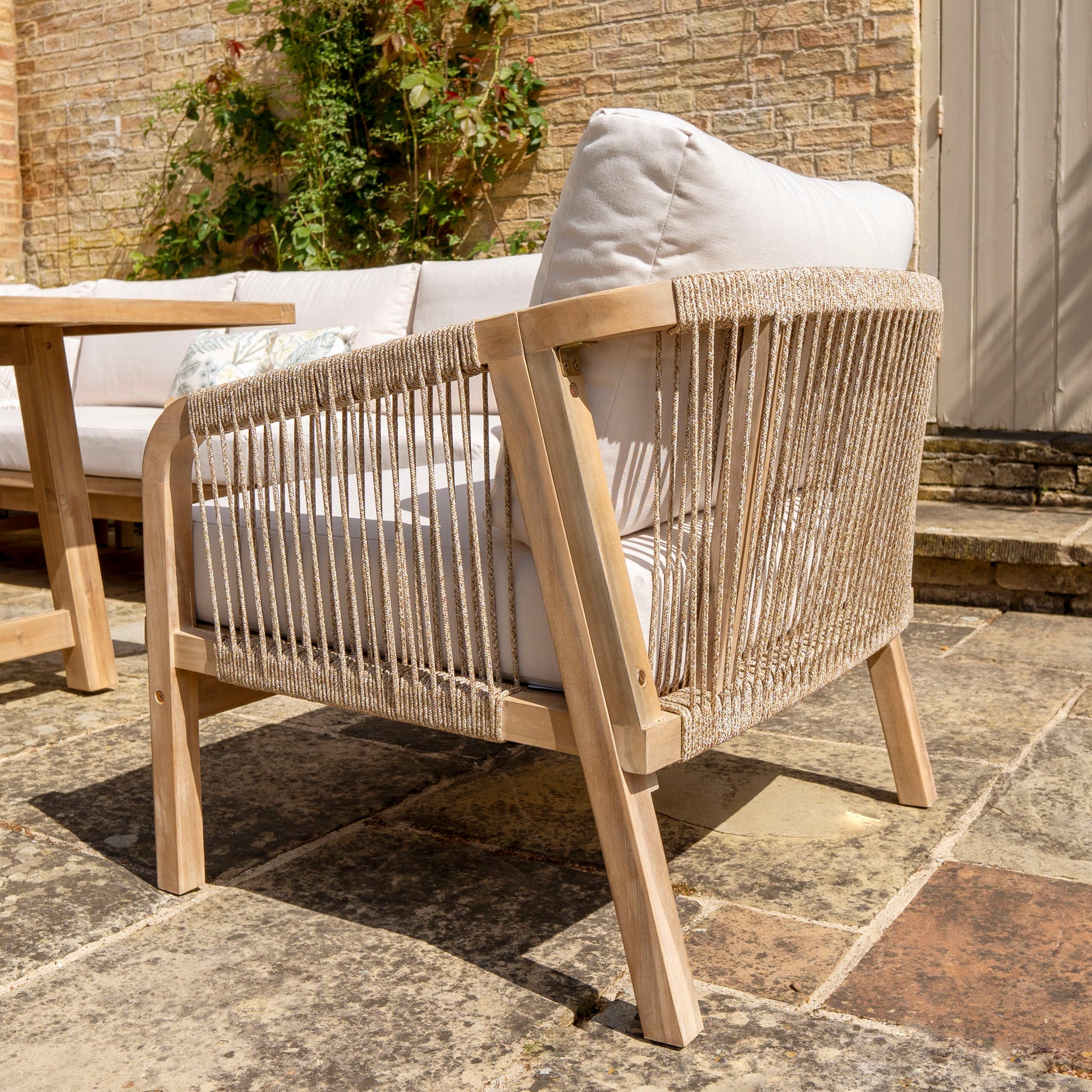 The Quay Corner Dining Set with Armchair in Linen, crafted from sustainably sourced acacia wood and featuring a cushioned seat and backrest, is elegantly placed on a stone patio near a brick wall adorned with climbing plants.