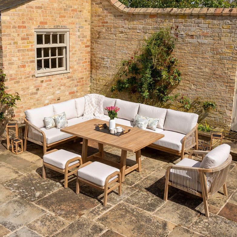 The Quay Corner Dining Set with Armchair in Linen showcases a wooden table made from sustainably sourced acacia wood, complemented by white cushioned seating, stools, and a decorative vase of pink flowers against a charming brick wall backdrop.