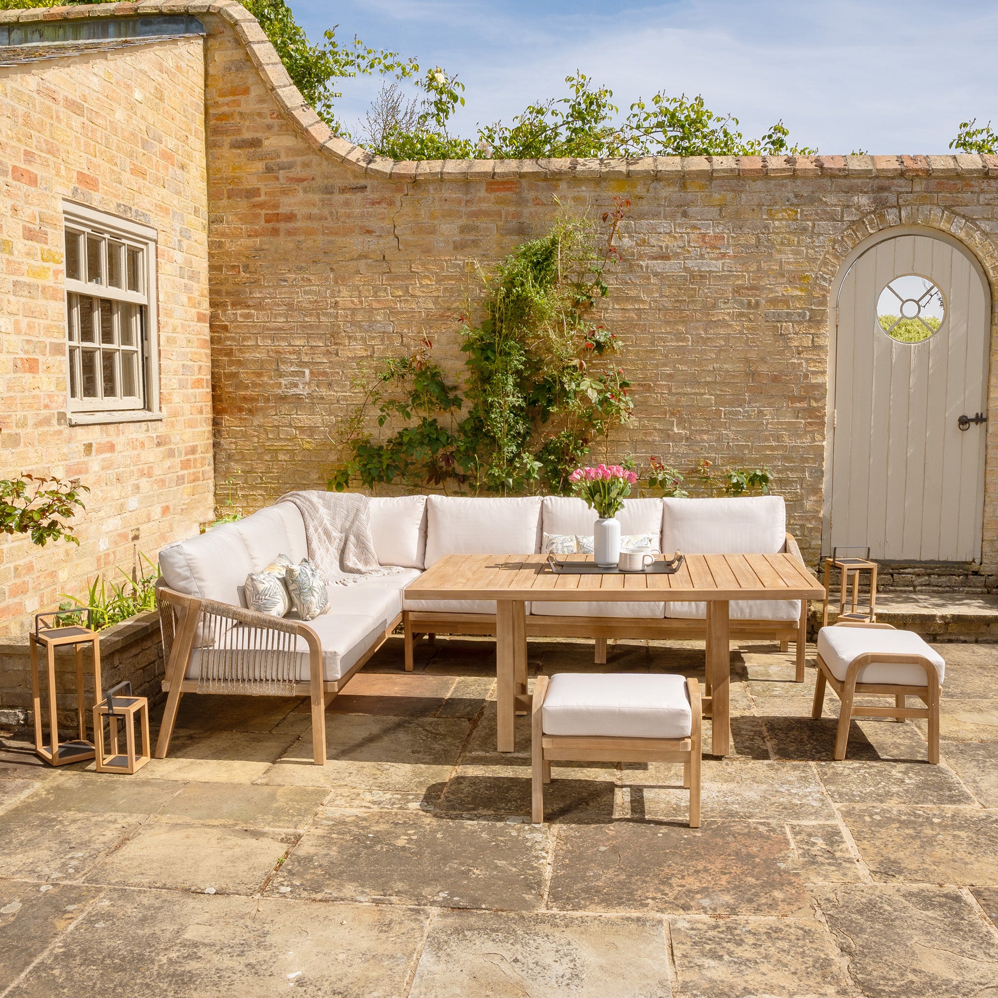 A cozy outdoor patio with the Quay Corner Dining Set in Linen, made of sustainably sourced acacia wood, featuring a cushioned corner sofa with water-resistant polyester fabric, a table, chairs, and garden decor.