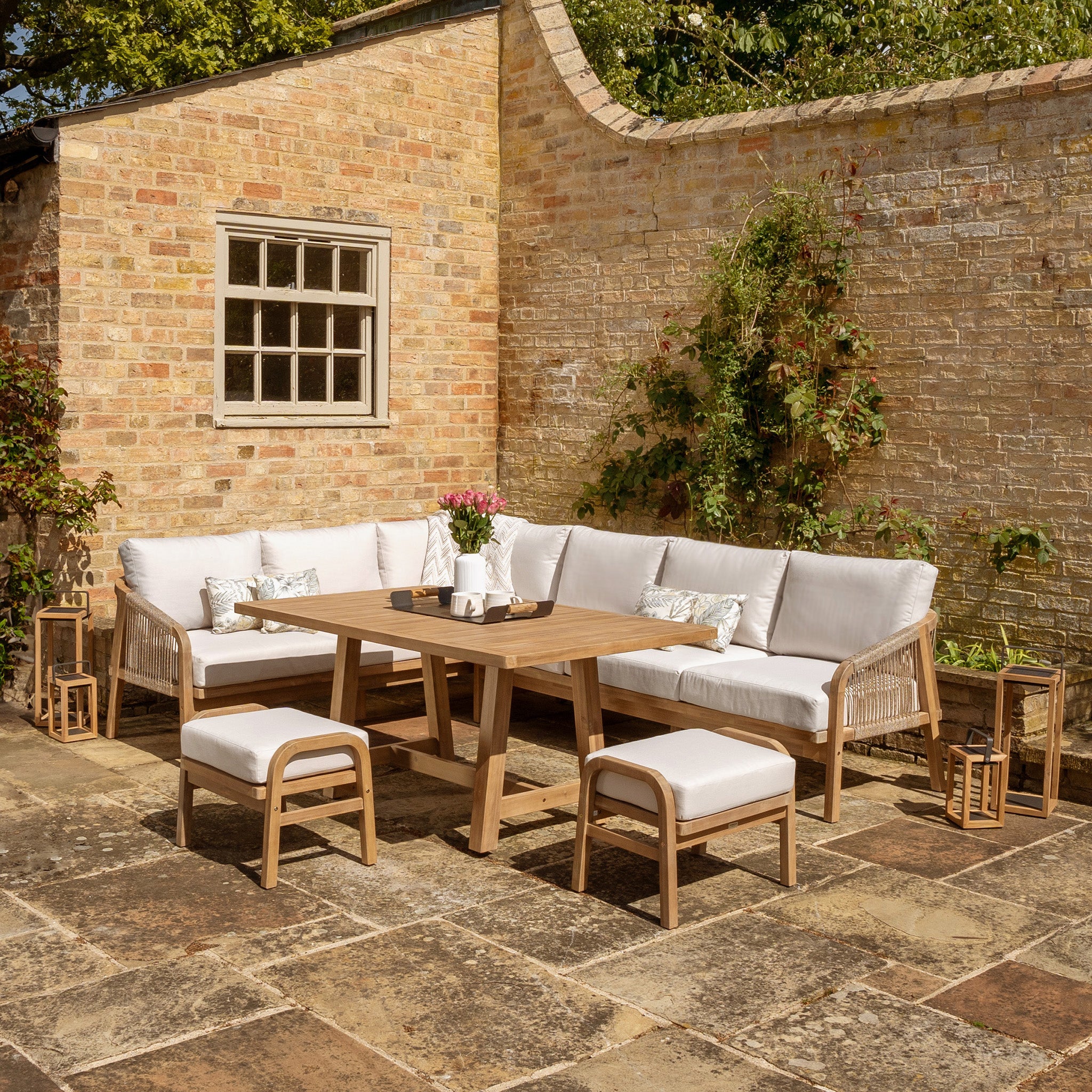 Outdoor patio with a wooden table made from sustainably sourced acacia wood, cushioned chairs with water-resistant polyester fabric, and a cozy corner sofa set against a brick wall. Part of the Quay Corner Dining Set in Linen.