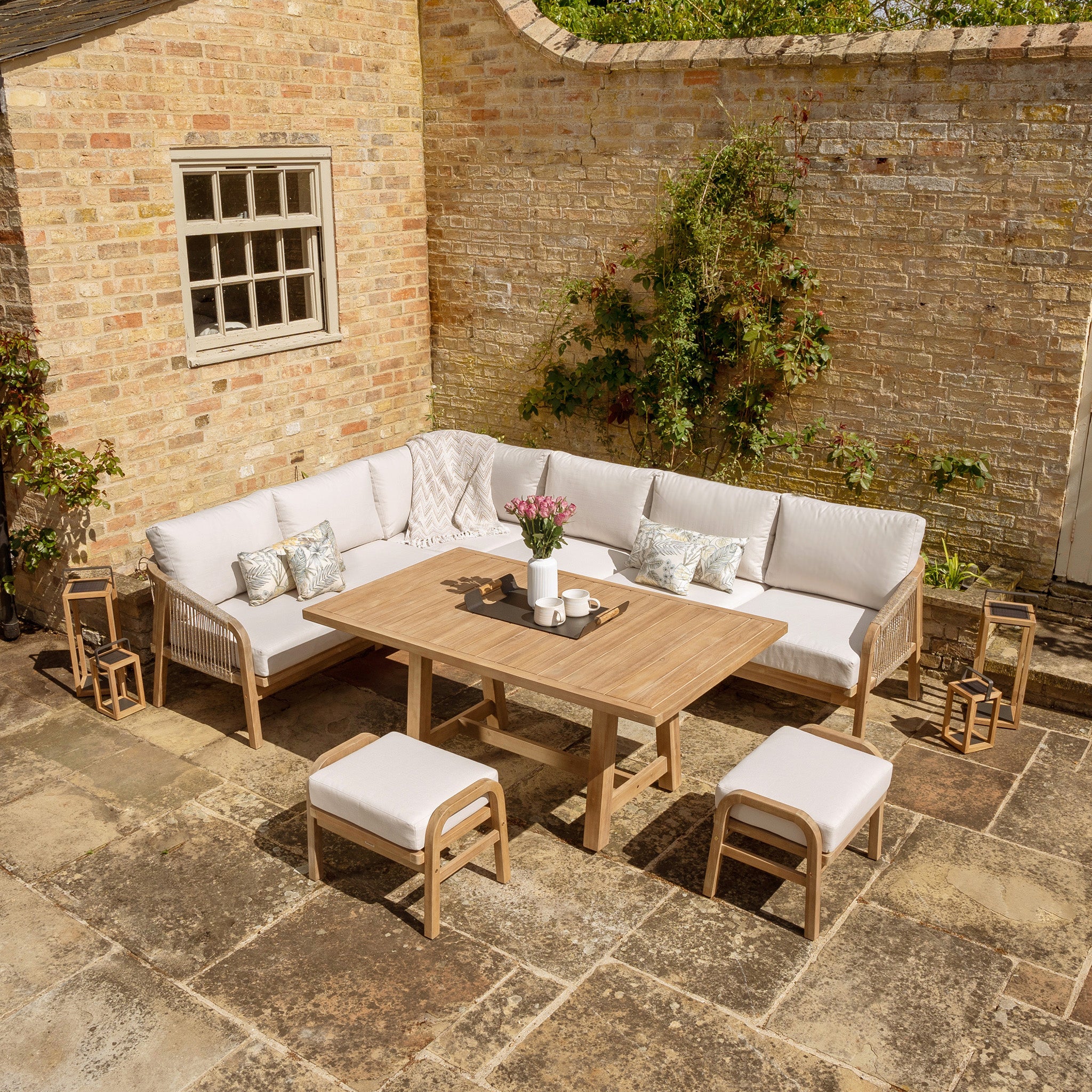 The Quay Corner Dining Set in Linen features an L-shaped sofa with beige cushions made of water-resistant polyester fabric, a dining table crafted from sustainably sourced acacia wood, matching stools, and lanterns, all set on stone tile flooring.