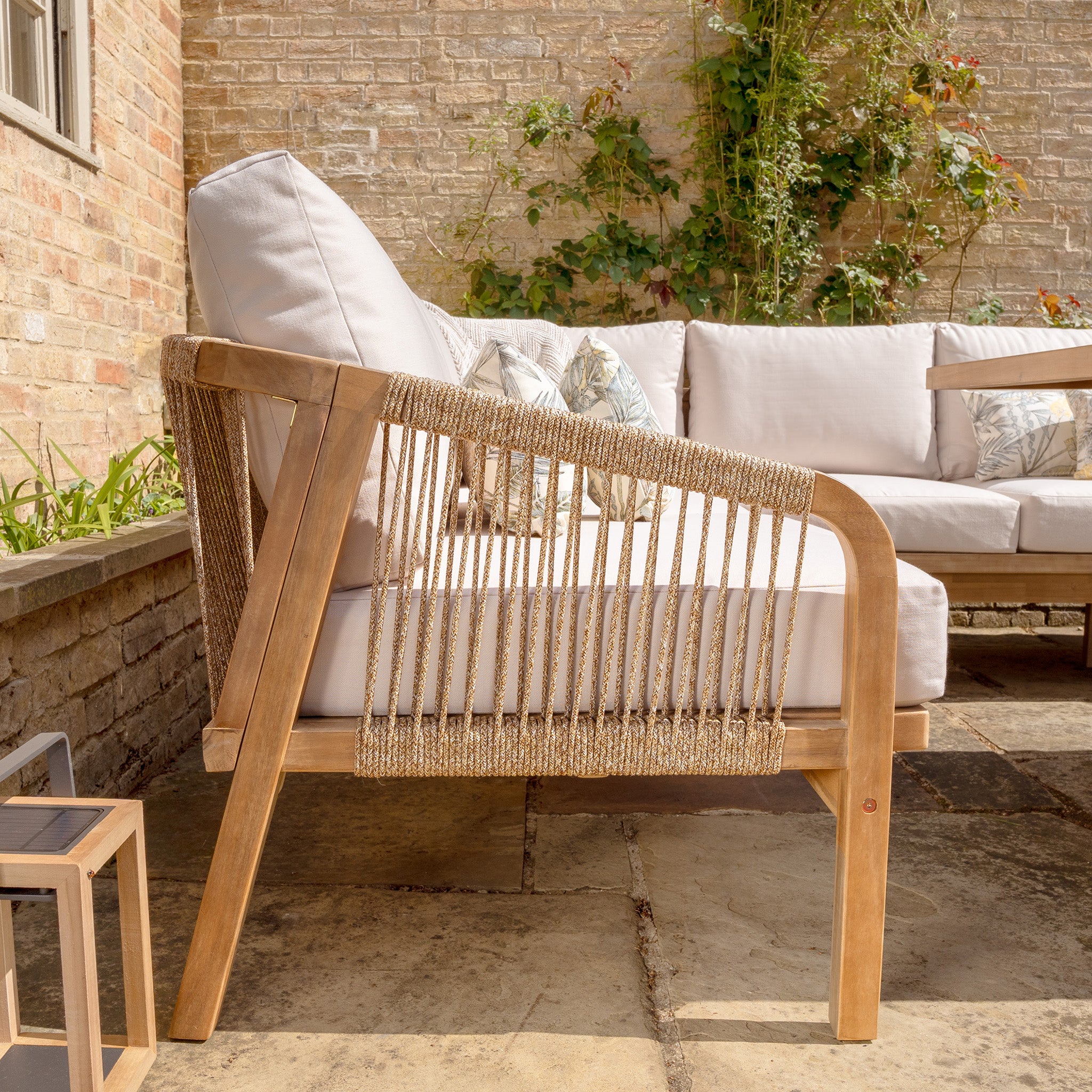 The Quay Corner Dining Set in Linen graces a stone patio against a brick wall adorned with climbing plants. The elegant setup features patio seating with wooden frames made from sustainably sourced acacia wood and beige cushions crafted from water-resistant polyester fabric.