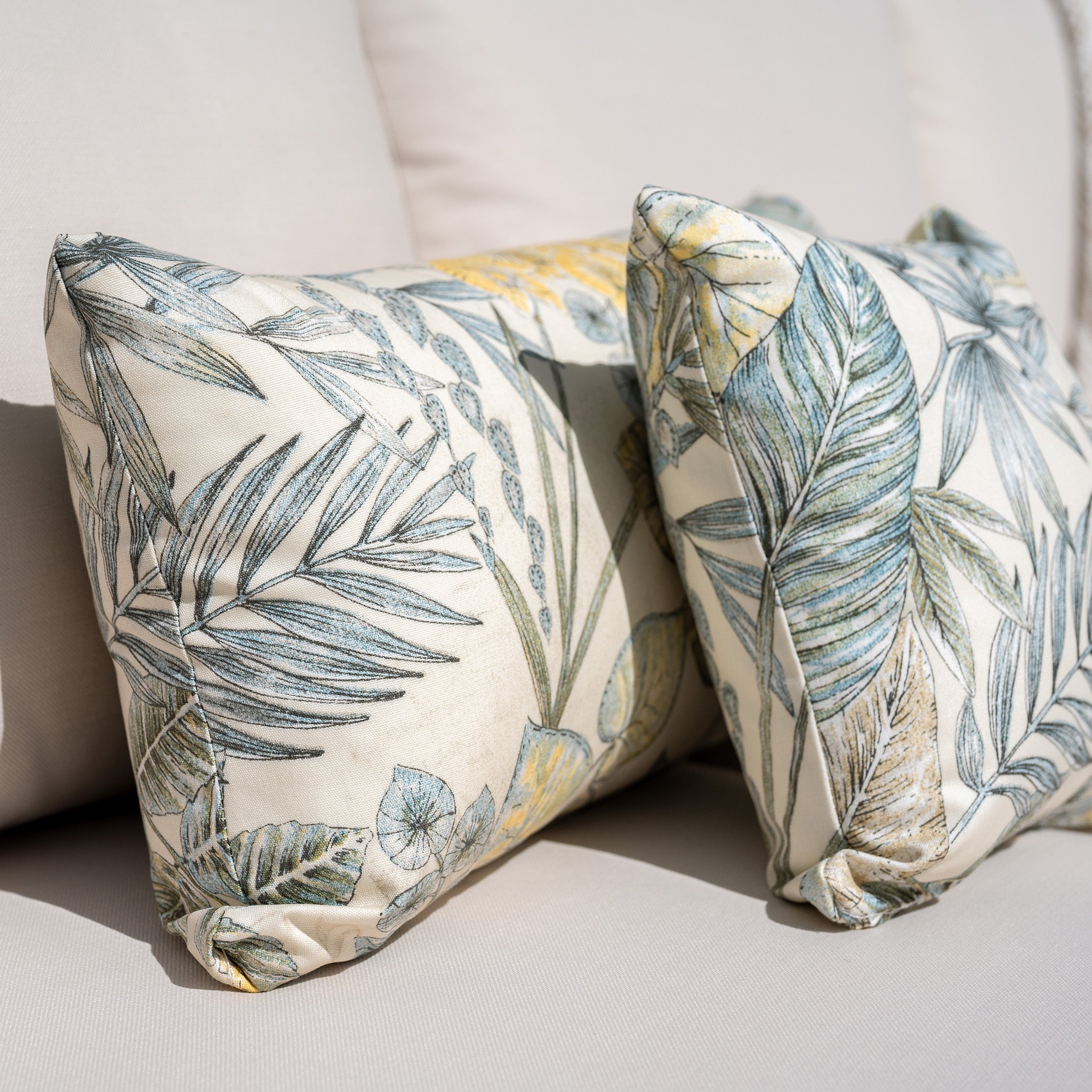 Two decorative pillows with a green and yellow tropical leaf print on a beige couch, adding a touch of tropical charm. Complement your living space with the Quay Corner Dining Set in Linen, crafted from sustainably sourced acacia wood for an eco-friendly and stylish ensemble.