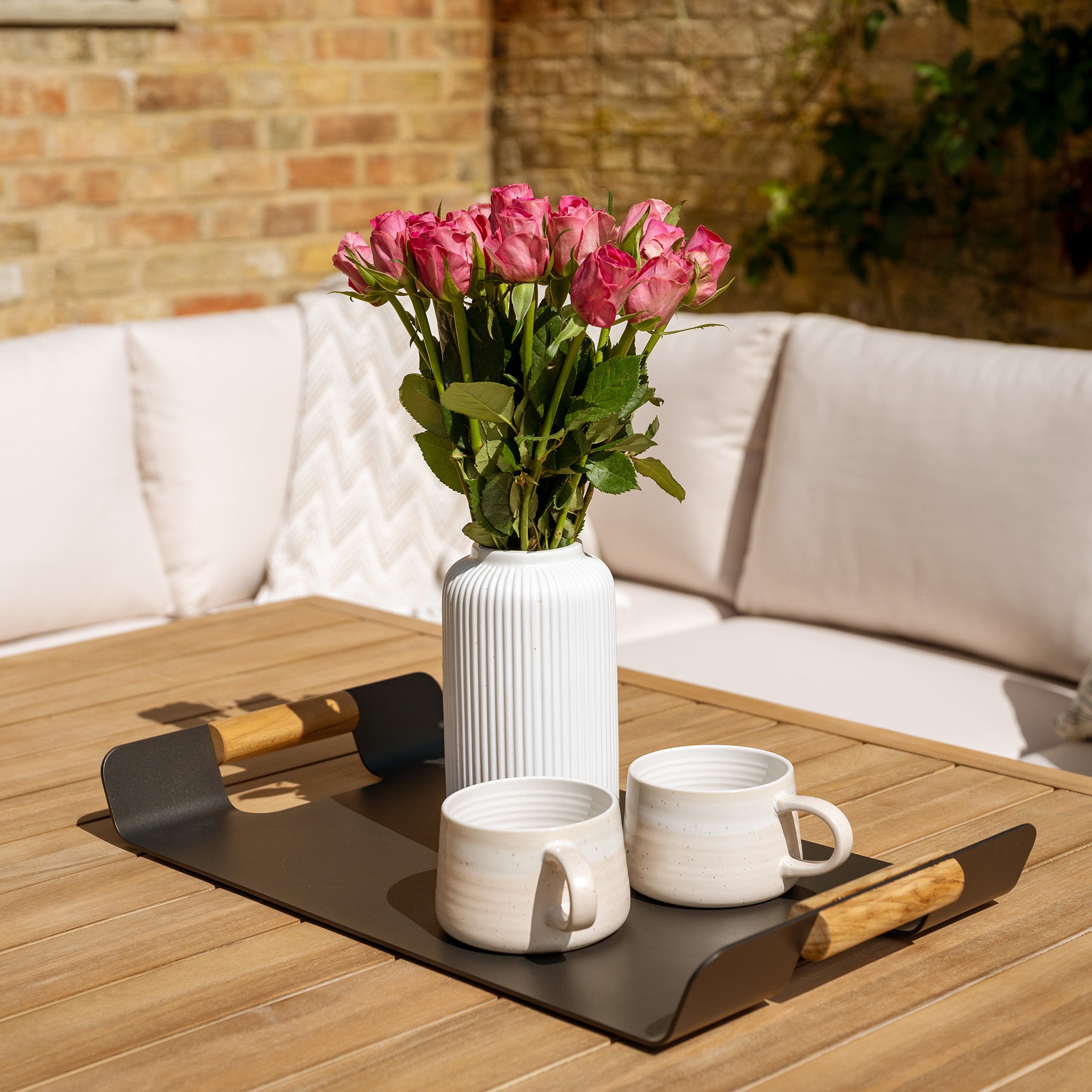 A vase of pink roses and two white mugs on a tray sit on a wooden table crafted from sustainably sourced acacia wood in a cozy outdoor seating area, part of the elegant Quay Corner Dining Set in Linen.