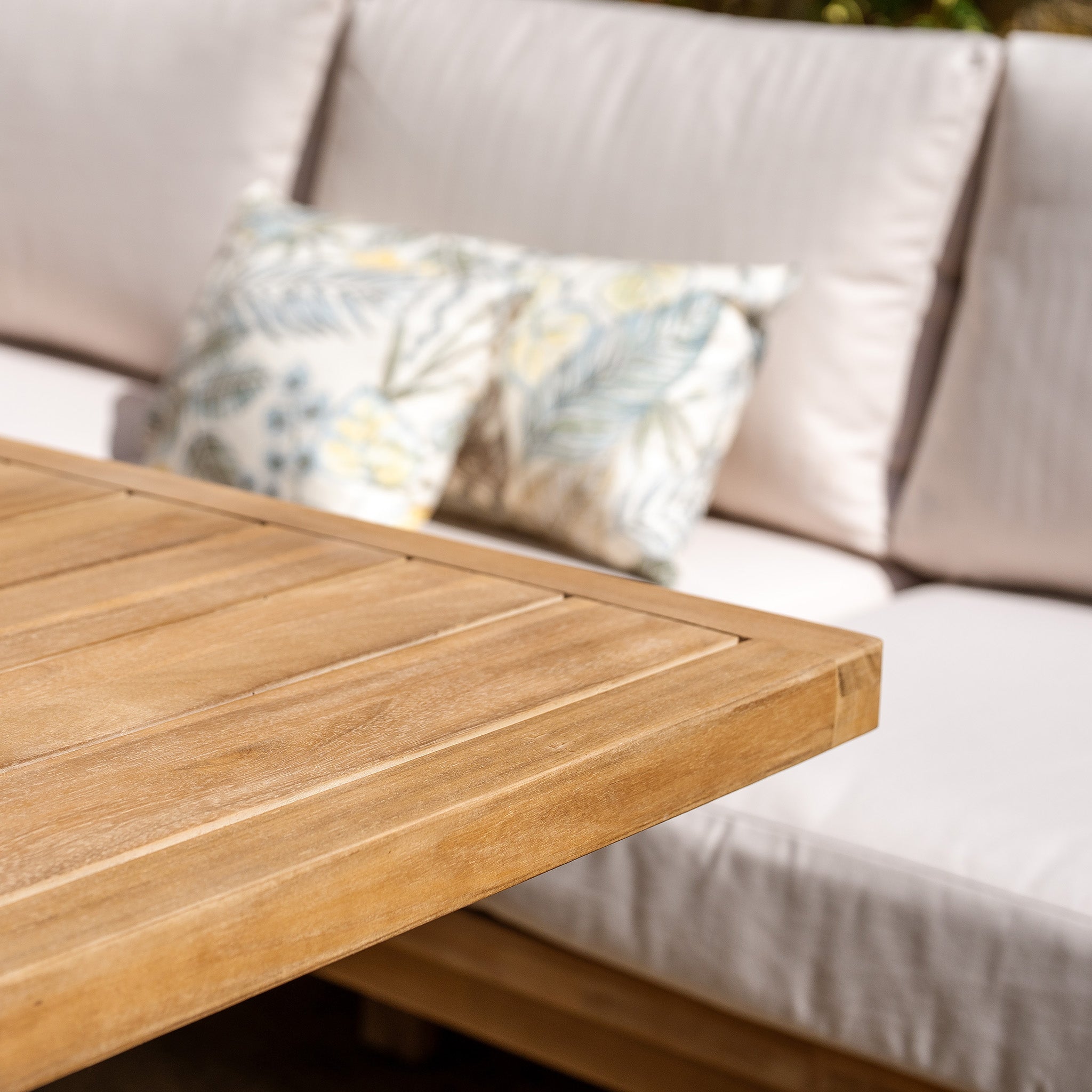 Close-up of the Quay Corner Dining Set in Linen, showcasing a wooden table made from sustainably sourced acacia wood, accompanied by a cushioned outdoor sofa and decorative pillows featuring water-resistant polyester fabric in the background.