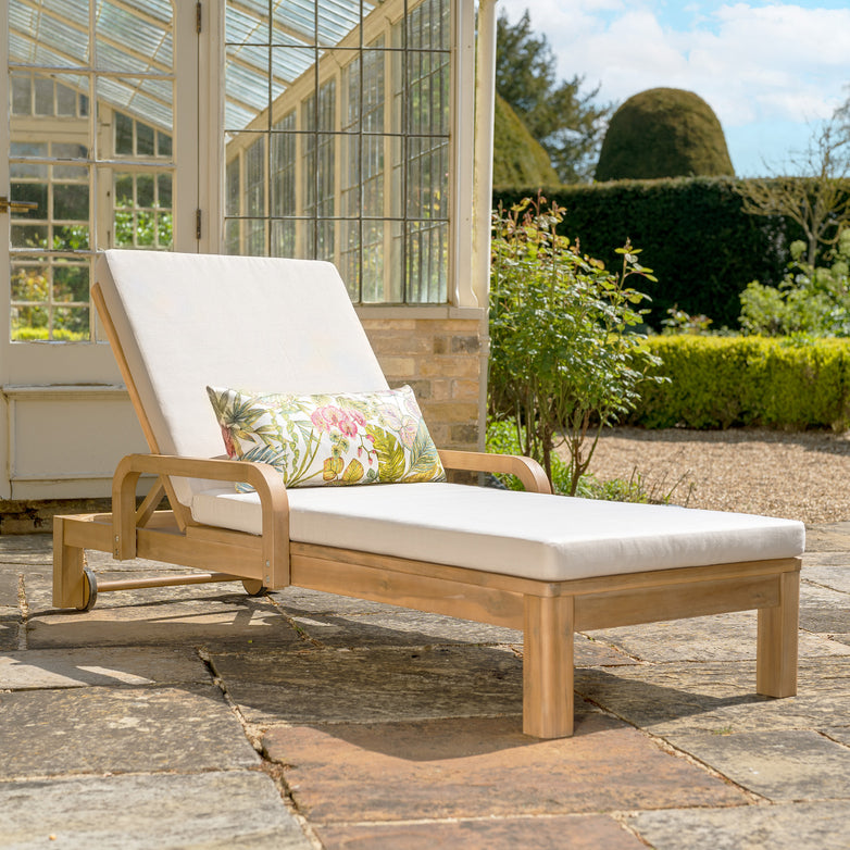 Part of the Quay collection, the Quay Sun Lounger in Linen is crafted from acacia wood and comes with white cushions and a floral pillow, positioned on a stone patio near a glass greenhouse on a sunny day.