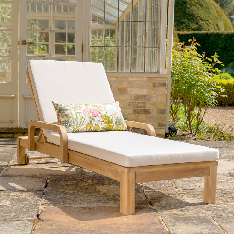 The Quay Sun Lounger in Linen, made from acacia wood, comes with white cushions and a floral pillow, making it an ideal addition to a stone patio near a glass-paneled garden room.