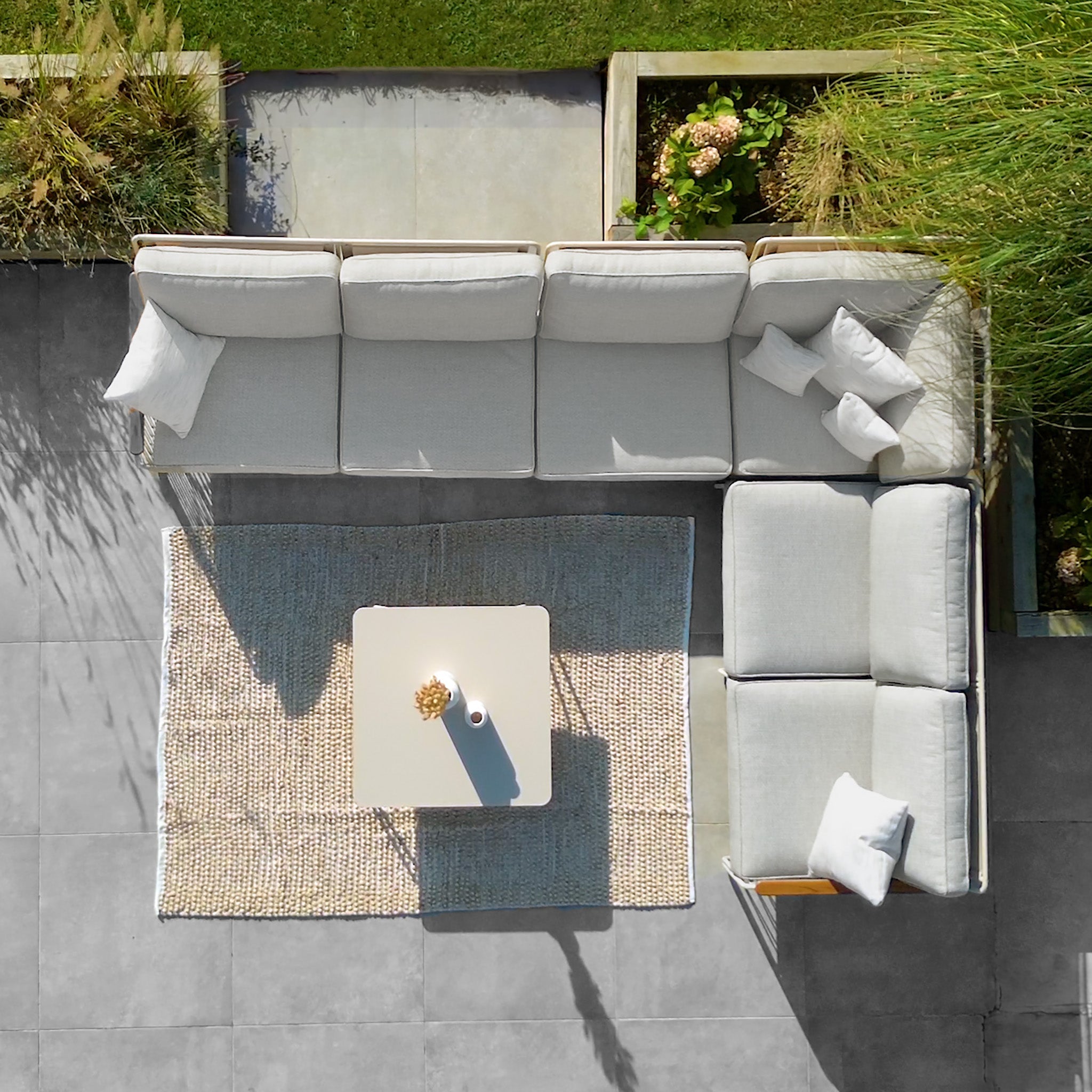 Aerial view of the Riviera Luxury Large Outdoor Corner Group Set in Latte, showcasing an L-shaped couch with hydrophobic rope weave and teak armrests, arranged around a small center table on a textured rug, surrounded by grass and plants.