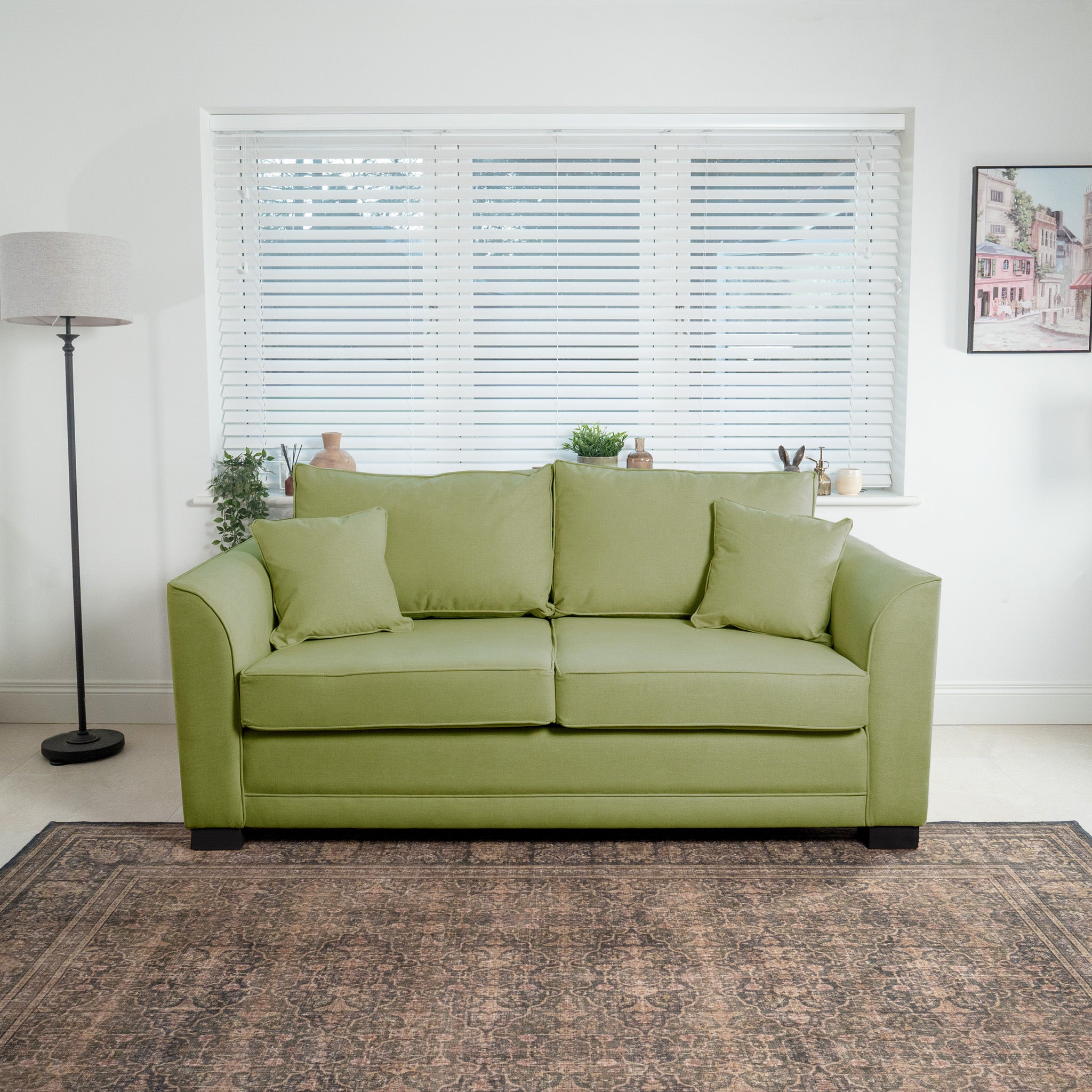 The Ardley 3 Seat Sofa Bed in olive, complete with cushions, sits in a bright room surrounded by plants and a floor lamp. With blinds covering the window, it adds a touch of luxury furniture elegance.