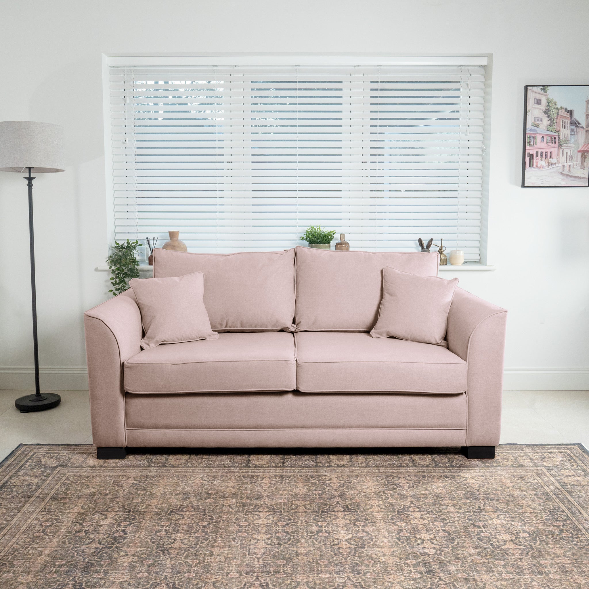 The Ardley 3 Seat Sofa Bed in Dusky Pink, featuring plush velvet and accompanying pillows, graces a bright room and is beautifully accented by a floor lamp, window blinds, and wall art. Everything rests elegantly on a patterned rug. This handcrafted UK piece brings sophistication and charm to any living space.