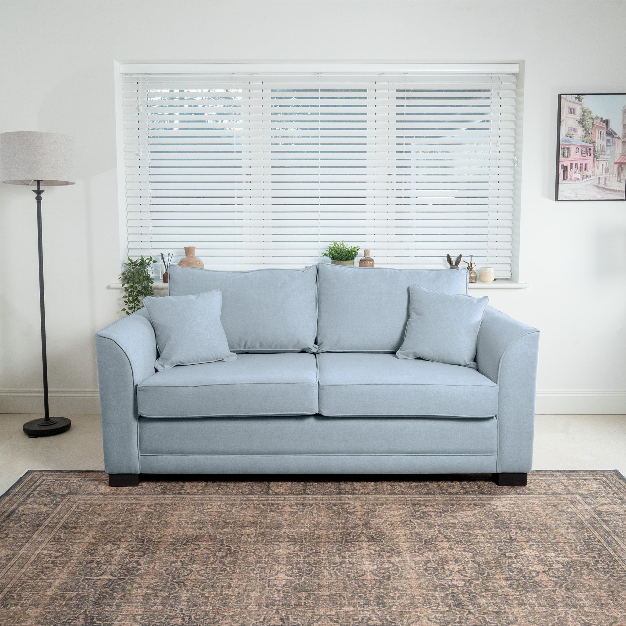 The Ardley 3 Seat Sofa Bed in sky blue is elegantly positioned in front of a window with blinds, enhanced by three cushions. A floor lamp stands nearby, providing a warm glow on the British-made furniture and the framed picture on the wall.