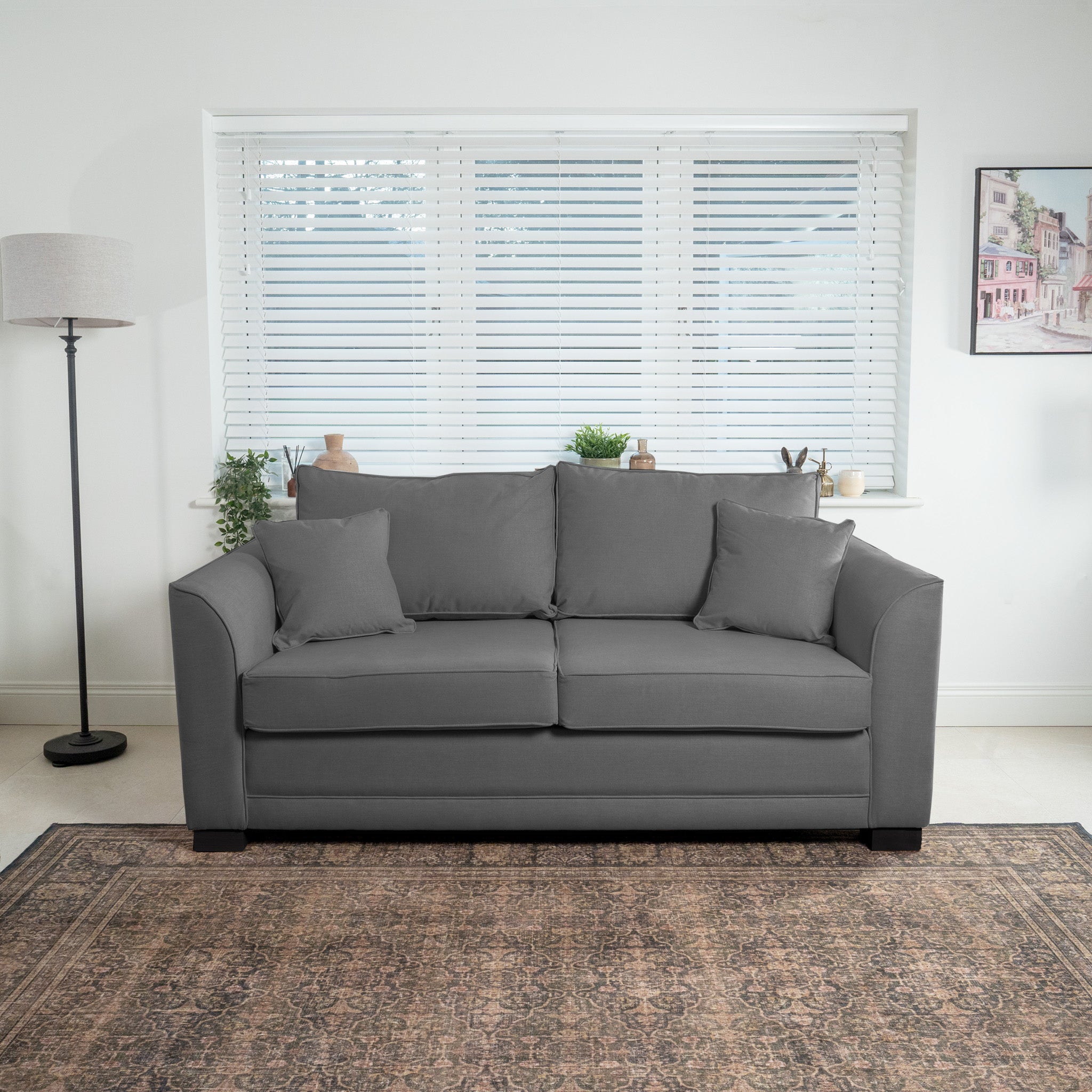 The Ardley 3 Seat Sofa Bed in Smoke Grey, featuring a pocket-sprung mattress and innovative Sedac mechanism for comfort, sits in front of a window with blinds. It is flanked by a floor lamp and potted plants, while a vintage rug graces the floor.