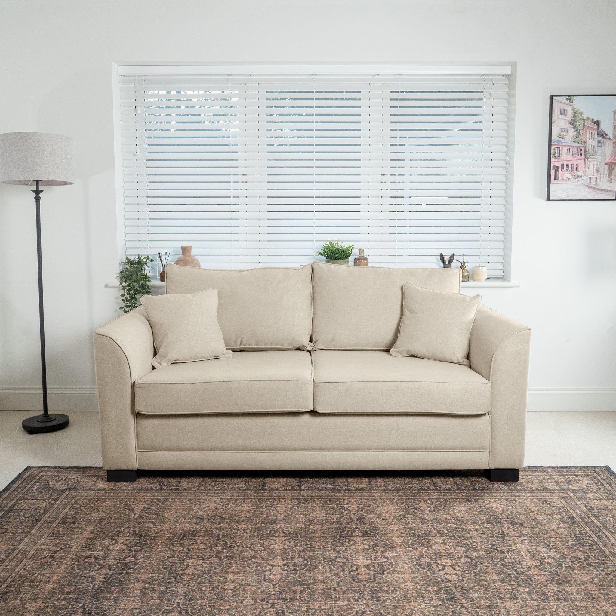 The Ardley 3 Seat Sofa Bed in Beige, crafted in Britain, graces the living room with its cushioned elegance. Complemented by a standing lamp and potted plants, it is positioned perfectly in front of a window adorned with blinds, offering both style and function.