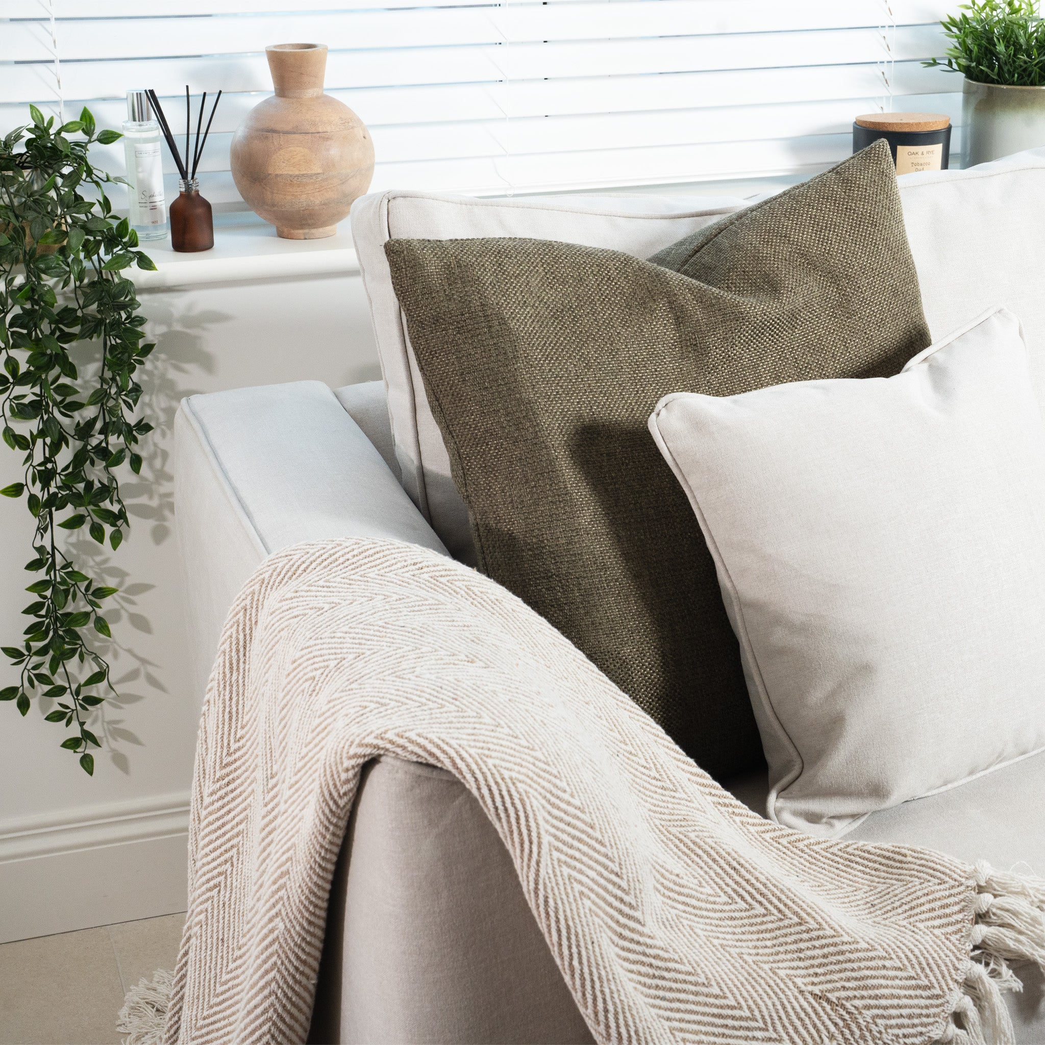 Discover the Ardley 3 Seat Sofa Bed, a British-made cozy piece in warm grey with beige cushions and a textured throw. It's nestled next to potted plants and decorative windowsill items, featuring a pocket-sprung mattress for ultimate comfort and functional charm.