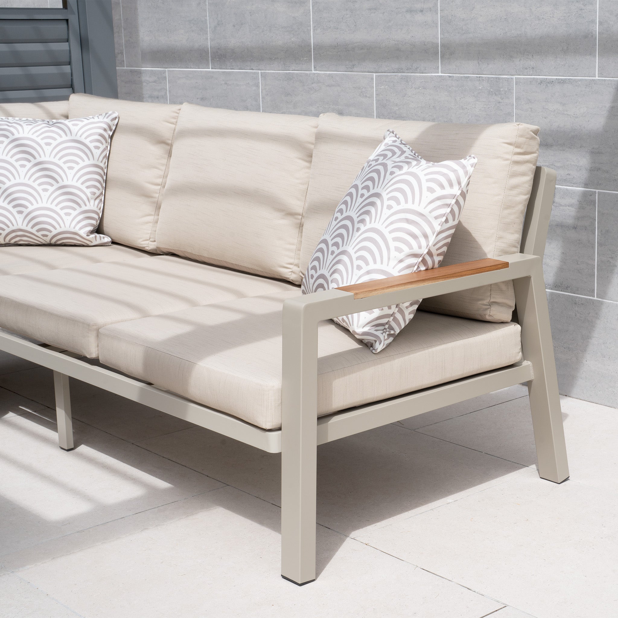 The Stockholm Corner Dining Set with Firepit Table in Limestone features a luxurious outdoor beige cushioned sofa adorned with white and geometric patterned pillows set against a tiled wall on a sunny day. This versatile garden furniture is perfect for complementing the firepit dining table, and its weatherproof cushions ensure lasting comfort and durability in any kind of weather.