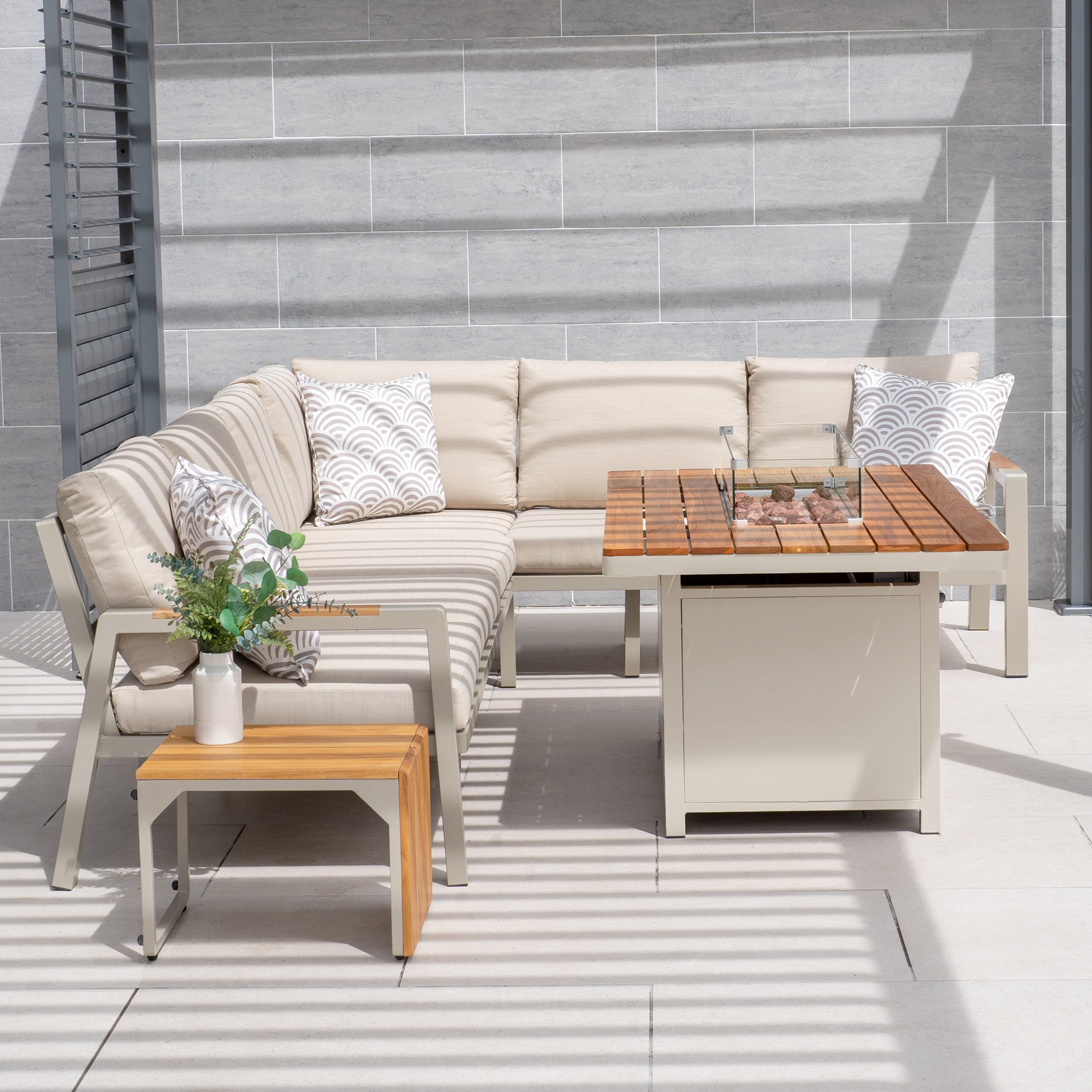 Outdoor seating area featuring the Stockholm Corner Dining Set with Firepit Table in Limestone, complete with weatherproof cushions, a wooden coffee table, a side table with a vibrant plant, and two patterned pillows.