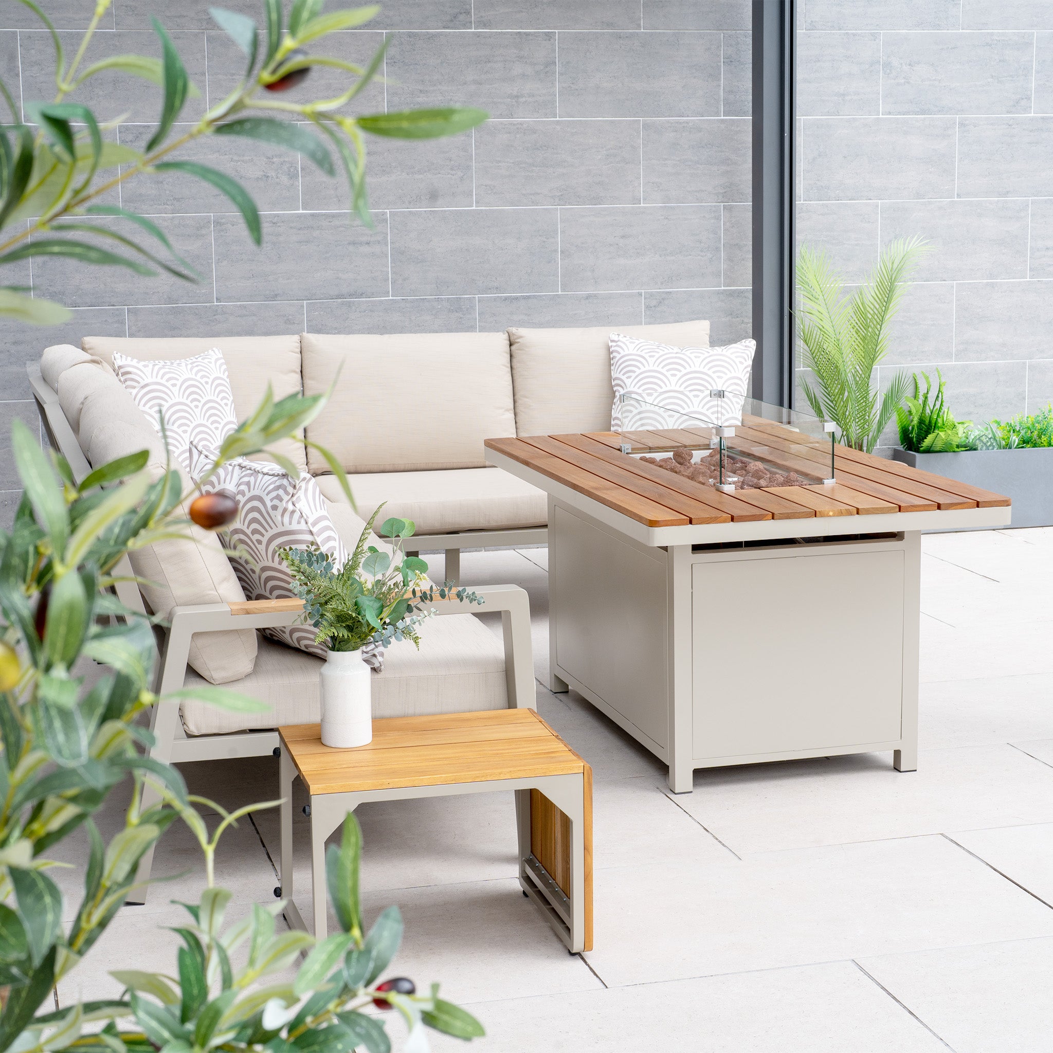 Outdoor patio featuring the Stockholm Corner Dining Set with Firepit Table in Limestone, complemented by a beige sectional sofa, weatherproof cushions, a small wooden side table, and lush plants.