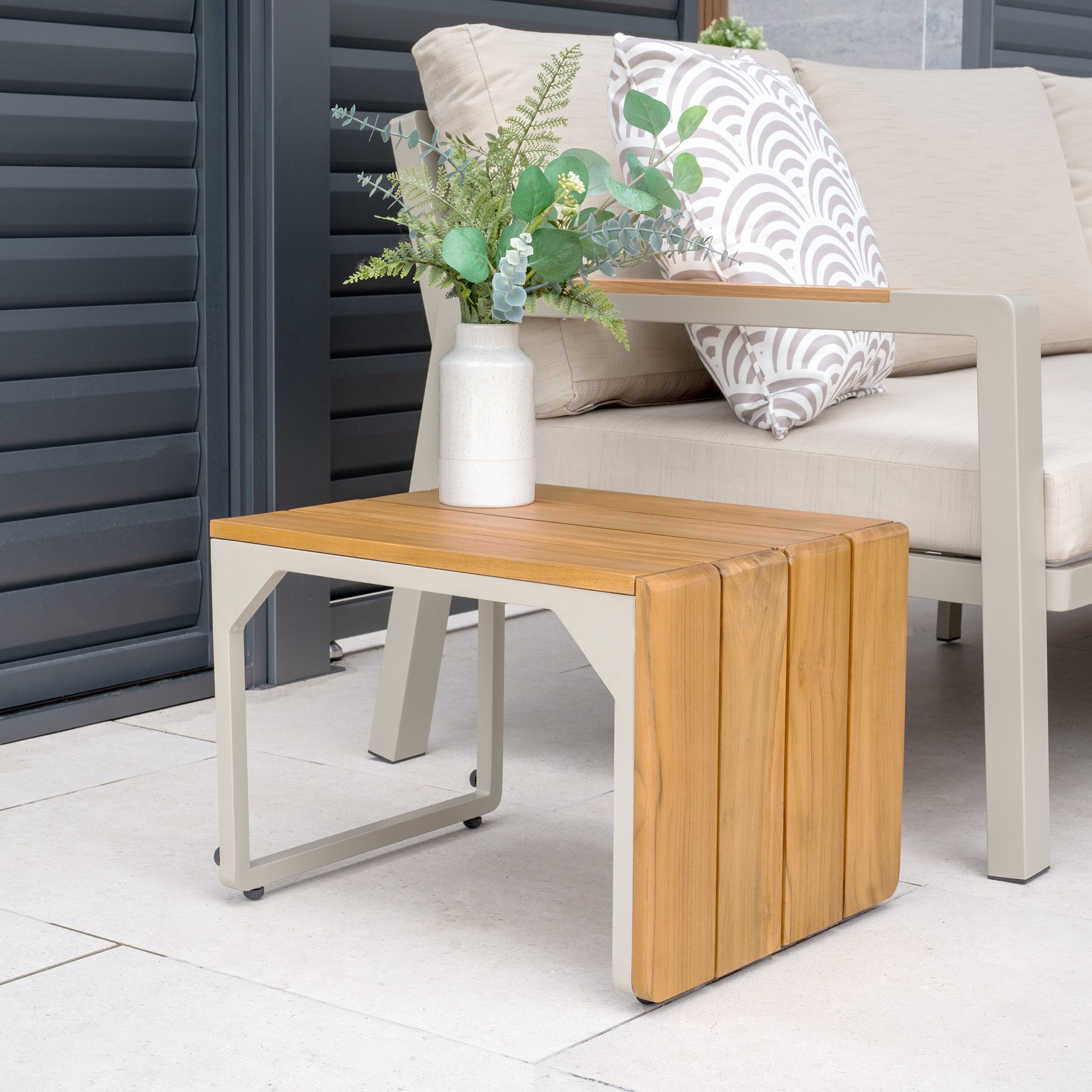 A teak side table with a white vase and plant complements a beige couch with patterned pillows, creating an inviting patio space. Nearby, the Pacific Lifestyle - Stockholm Corner Dining Set with Firepit Table in Limestone adds elegance for gatherings.