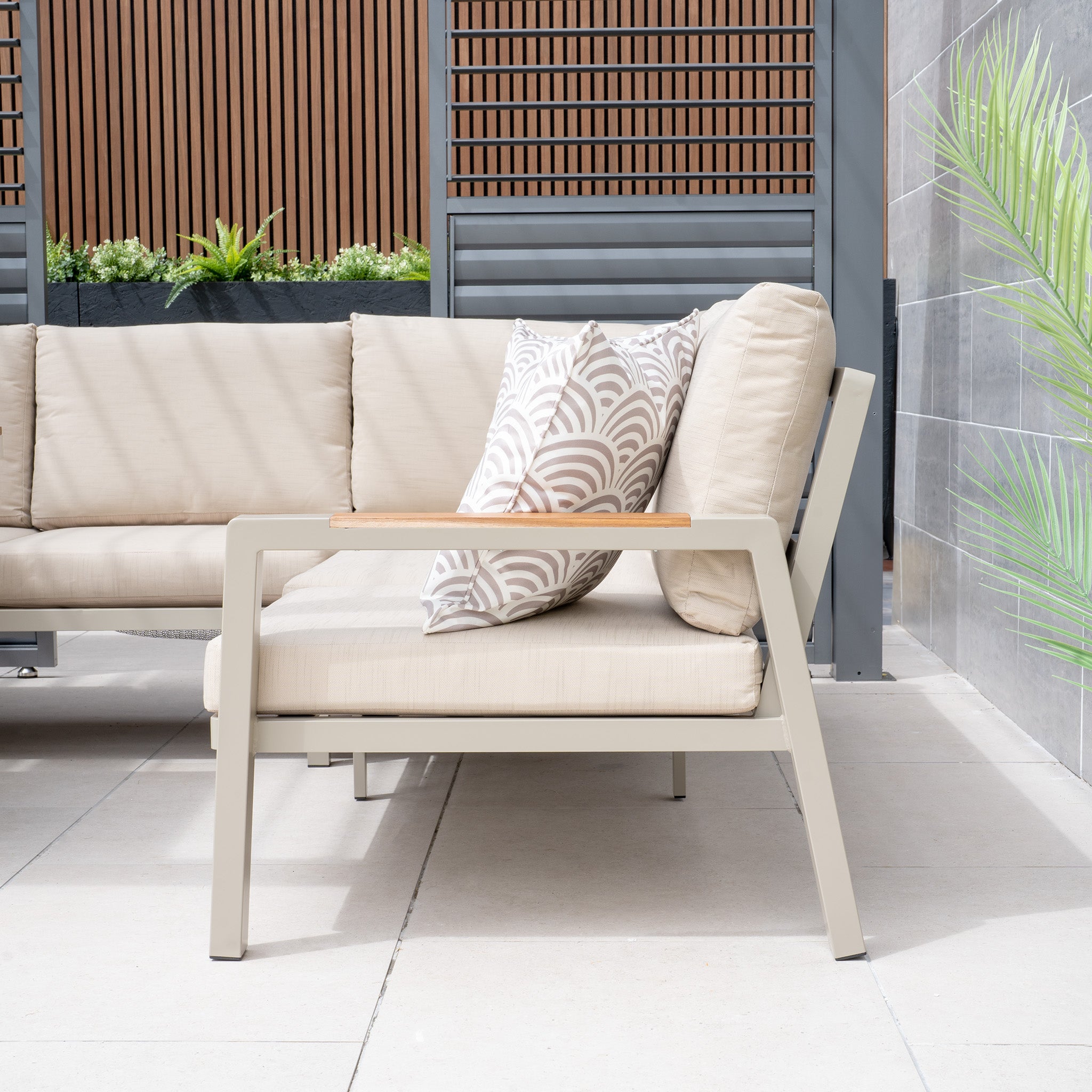 The beige patio sofa features patterned, weatherproof cushions and is set on a tiled floor with a wooden screen and potted plants in the background. It's an ideal garden furniture arrangement completed with the Stockholm Corner Dining Set with Firepit Table in Limestone for cozy gatherings.