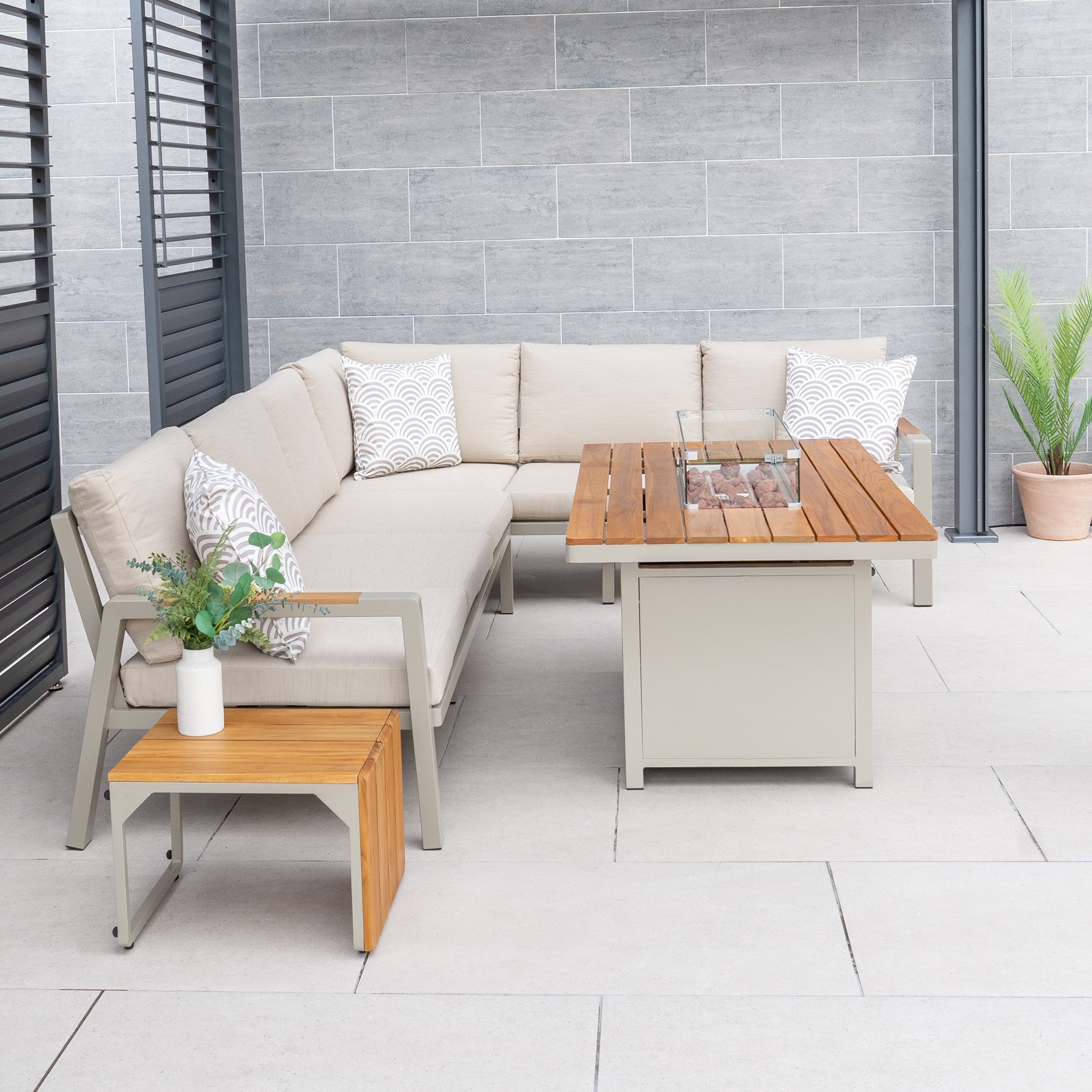 Modern patio featuring the Stockholm Corner Dining Set with Firepit Table in Limestone, complemented by a wooden table, a side table adorned with plants, and gray tile flooring. The garden furniture includes weatherproof cushions for year-round comfort.