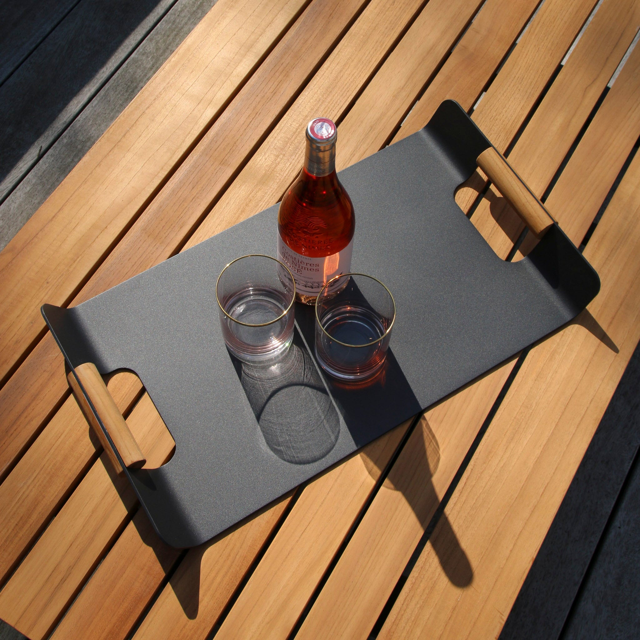 A Tracy Serving Tray with Teak Handles in Charcoal, holding a bottle of rose wine and two empty glasses, sits elegantly on a wooden table, complementing the natural beauty of the garden furniture.