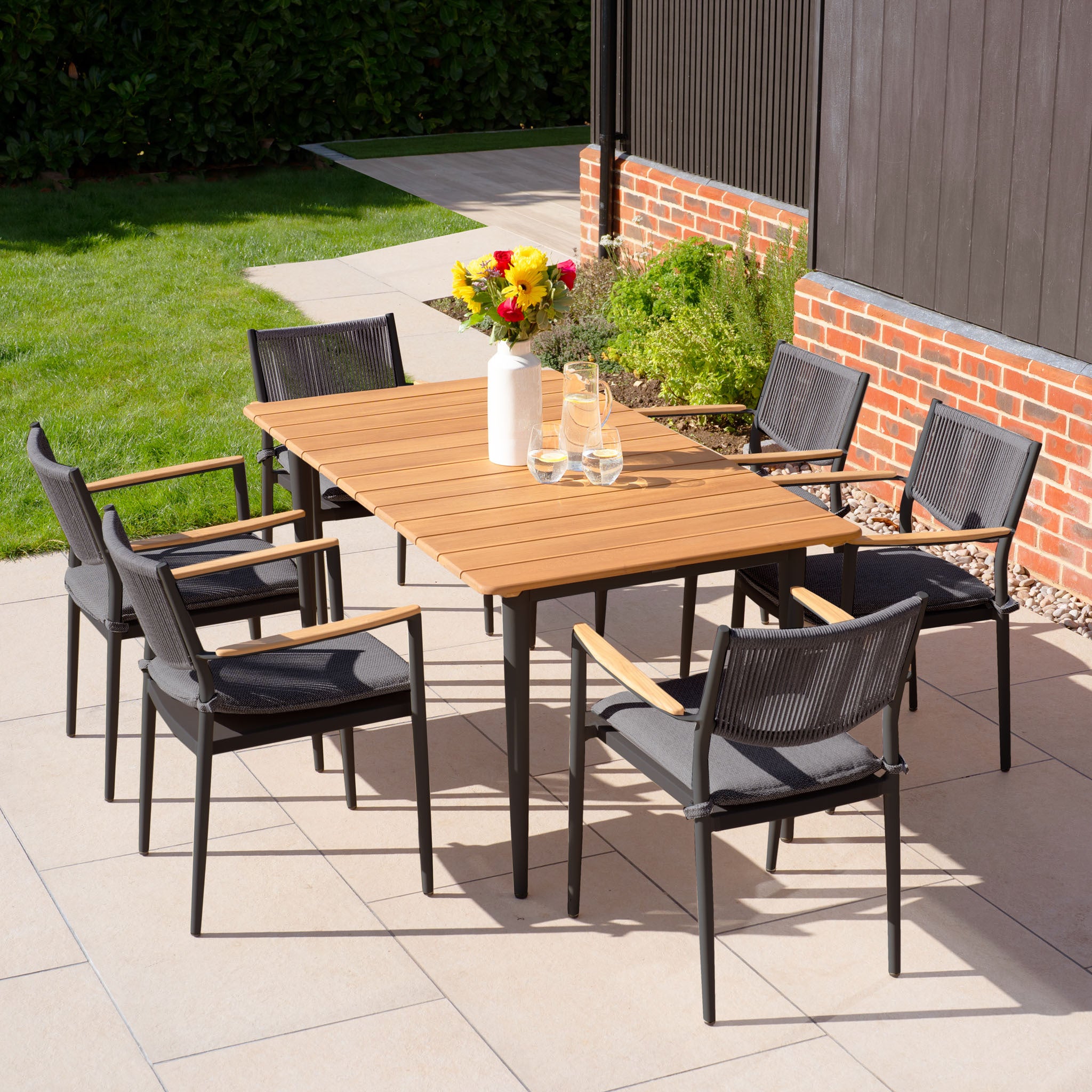 The patio features the Tellaro 6 Seat Rectangular Dining Set with a teak table in charcoal, paired with a charming vase of flowers for an elegant outdoor setup.