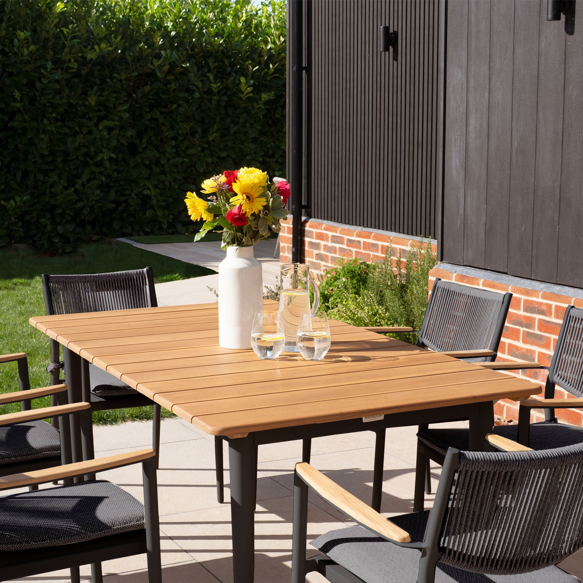 A Tellaro 6 Seat Rectangular Dining Set with a teak table in charcoal, complete with chairs, a vase of flowers, and glasses of water, is elegantly arranged outside the building.